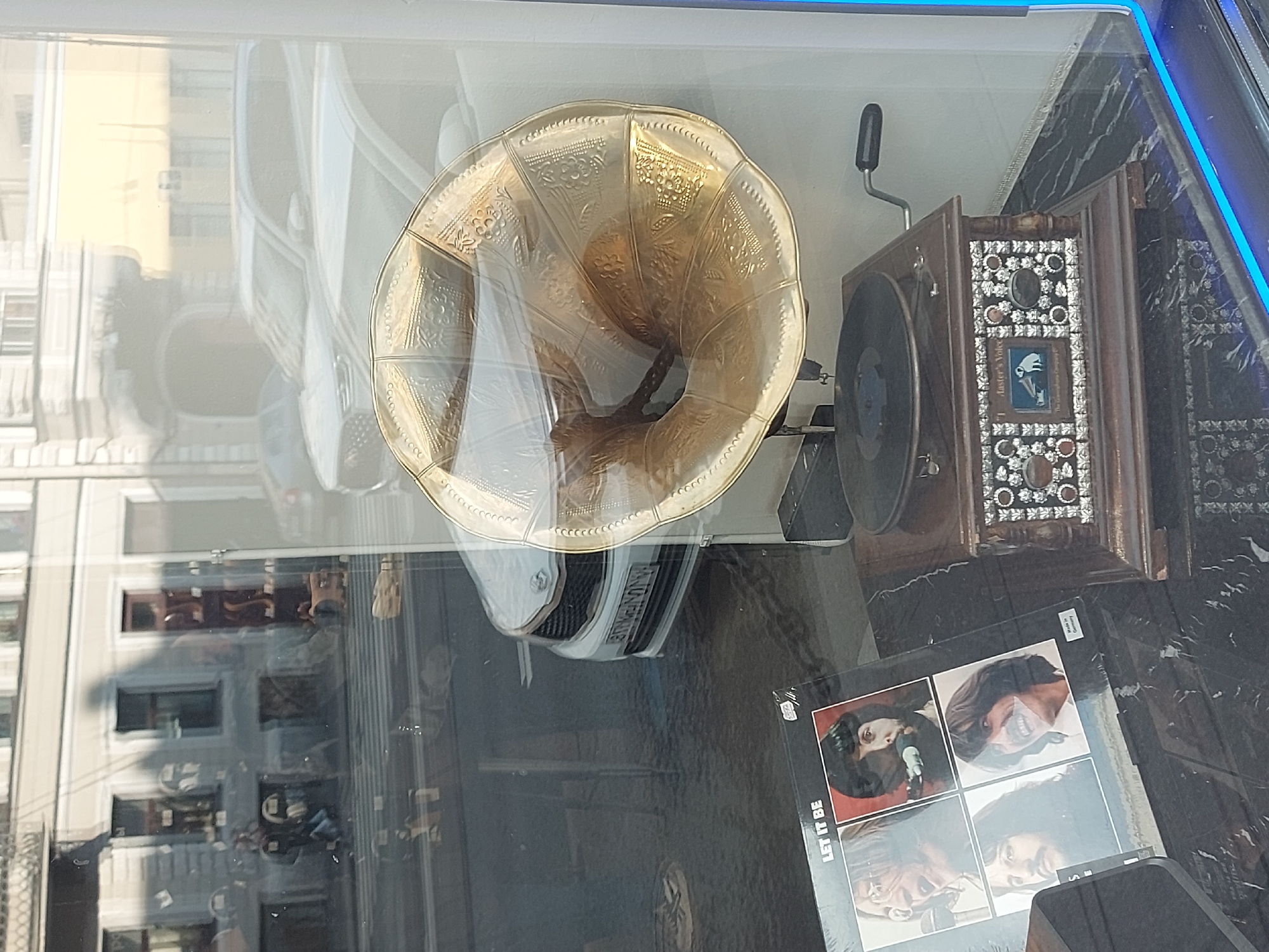 The image appears to be taken through a window in Saint Petersburg, Russia. The reflection in the window shows a street scene with pedestrians, buildings, and cars. Inside, there's an antique gramophone with a large, ornate brass horn and a record player component. A record album cover, "Let It Be" by The Beatles, is nearby. The window itself may be part of a display case in a store, possibly an antique shop.