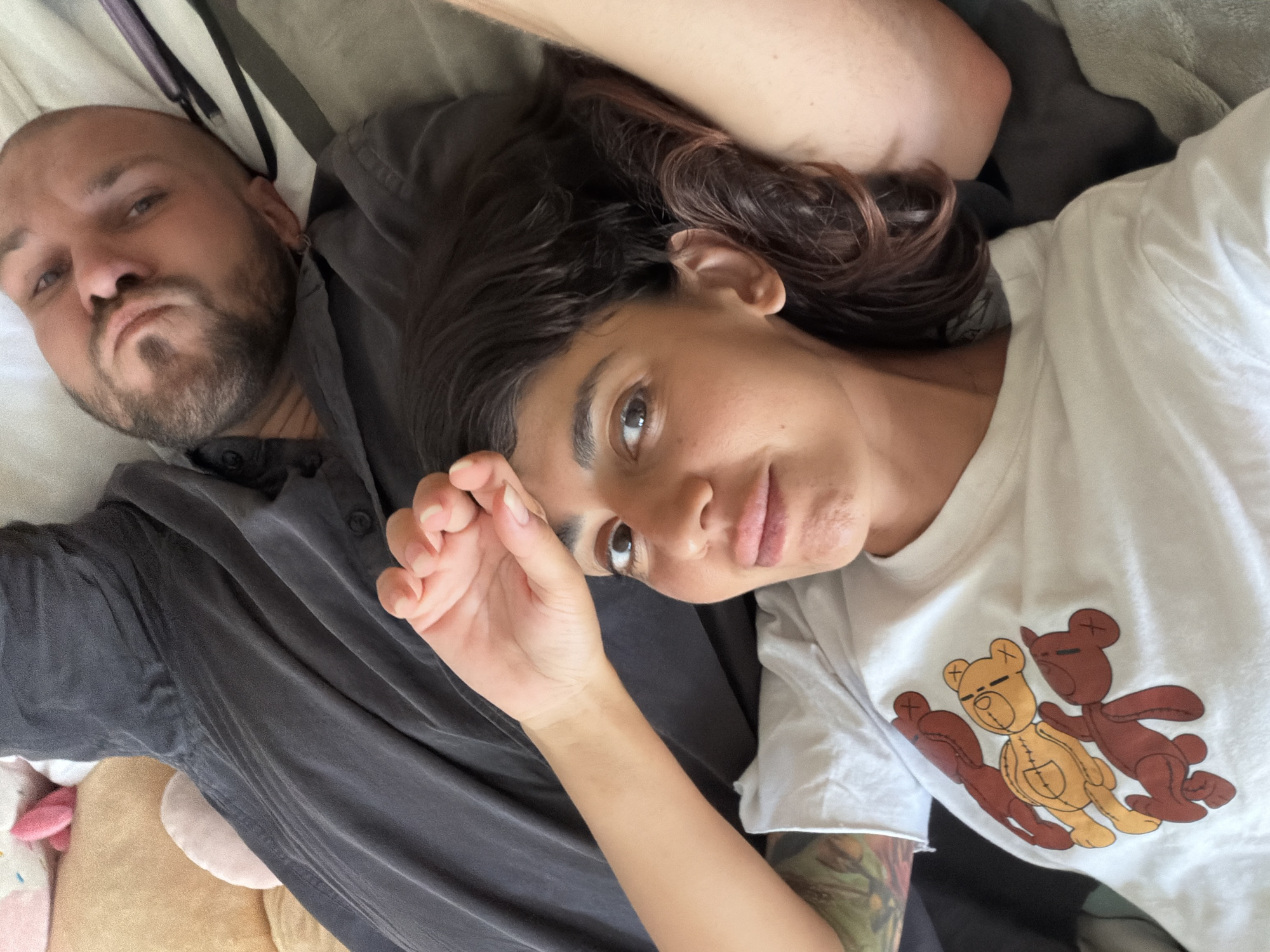 A close-up, high-angle shot shows a couple lying down in what appears to be a bed in Kuta Selatan, Indonesia. The man, with a shaved head and beard, is in the background, looking upwards with his eyes slightly closed. He is wearing a dark gray shirt. The woman is in the foreground, her head resting on his chest, looking at the camera with her right hand resting on her forehead. She has dark, wavy hair and is wearing a white t-shirt with a graphic of three teddy bears. The environment appears to be indoors, with soft lighting. There are stuffed animals visible in the upper left corner of the image, suggesting a bedroom setting.