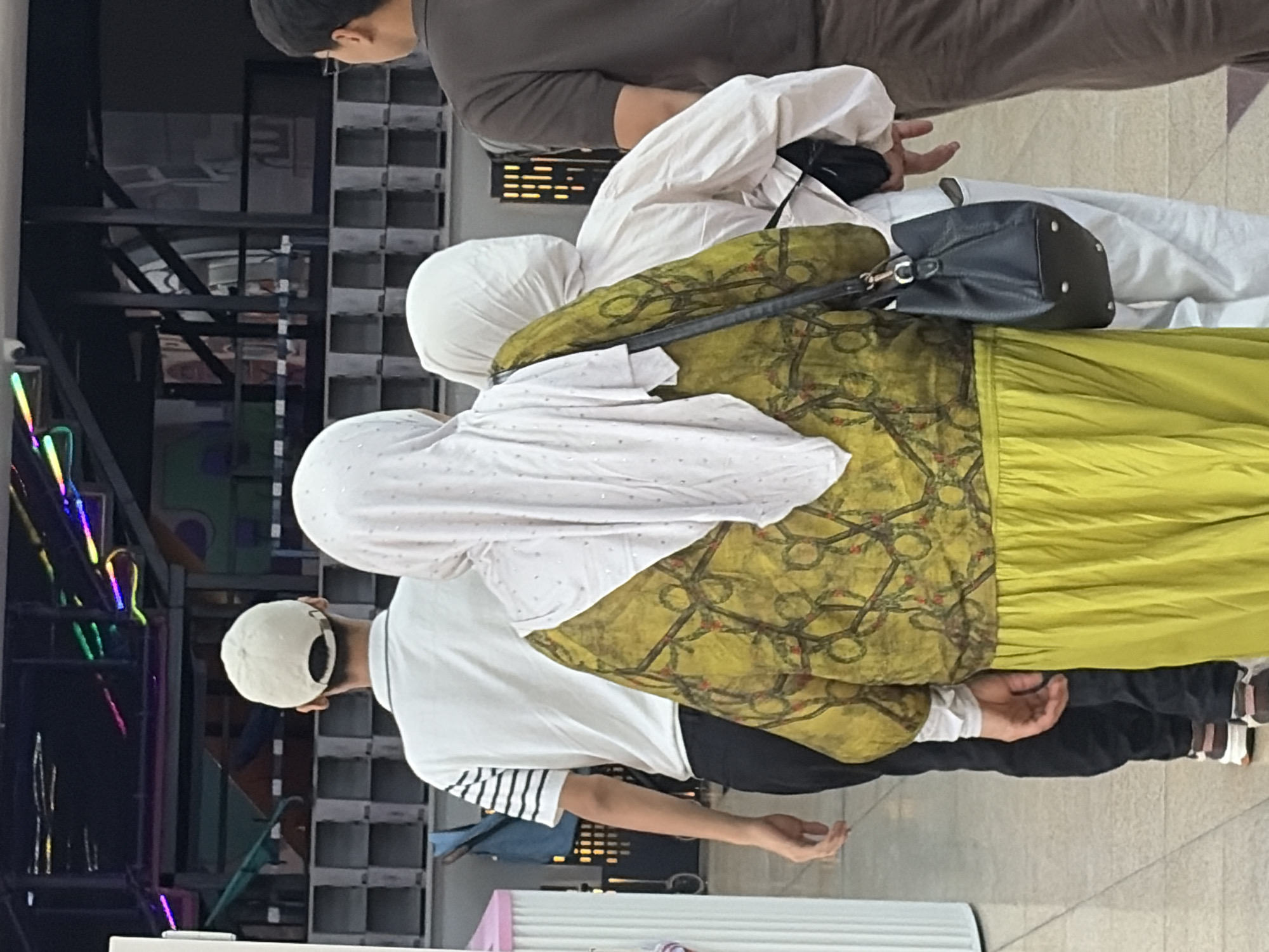 The image shows a group of people walking in a brightly lit indoor space. In the foreground, a woman wearing a green patterned tunic over a lime green skirt and a white headscarf is walking away from the camera. She is carrying a black handbag. Next to her, a person wearing a white outfit and a white headscarf is walking in the same direction. To the left of the woman in the green tunic, a person wearing a white t-shirt with black stripes on the sleeves and a white baseball cap is walking away from the camera. Further to the right, a person in a brown shirt is also walking away. The background shows a structure with colorful lights and rows of cubicle-like openings. The floor is tiled.