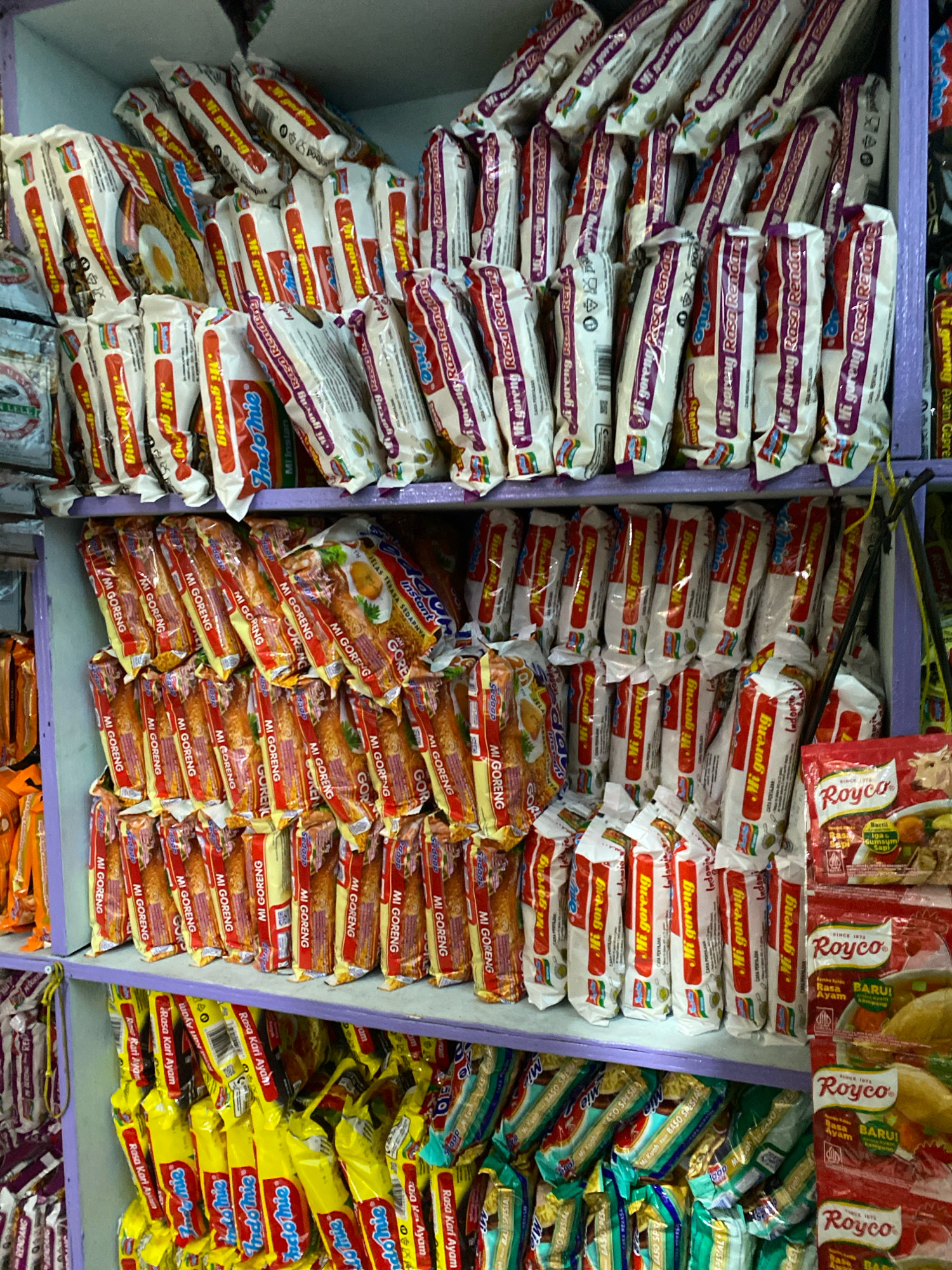 The image shows a close-up, eye-level view of shelves stocked with packages of instant noodles and soup seasoning in a store. The shelves are purple and appear to be in a dimly lit environment. The packaging of the products is predominantly red, white, and yellow, with some purple and green elements. Many of the packages are branded with Indonesian text, indicating local products. Specifically, "Indomie" and "Royco" are clearly visible, along with text such as "Mi Goreng," "Rasa Rendang," "Rasa Kari Ayam," and "Bumbu Kaldu Ayam Kampung." The sheer volume and arrangement suggest a retail setting, likely a small shop or minimarket common in Indonesian cities. Based on the prominent display of these popular Indonesian brands, the scene is consistent with a retail environment in Semarang, Indonesia.