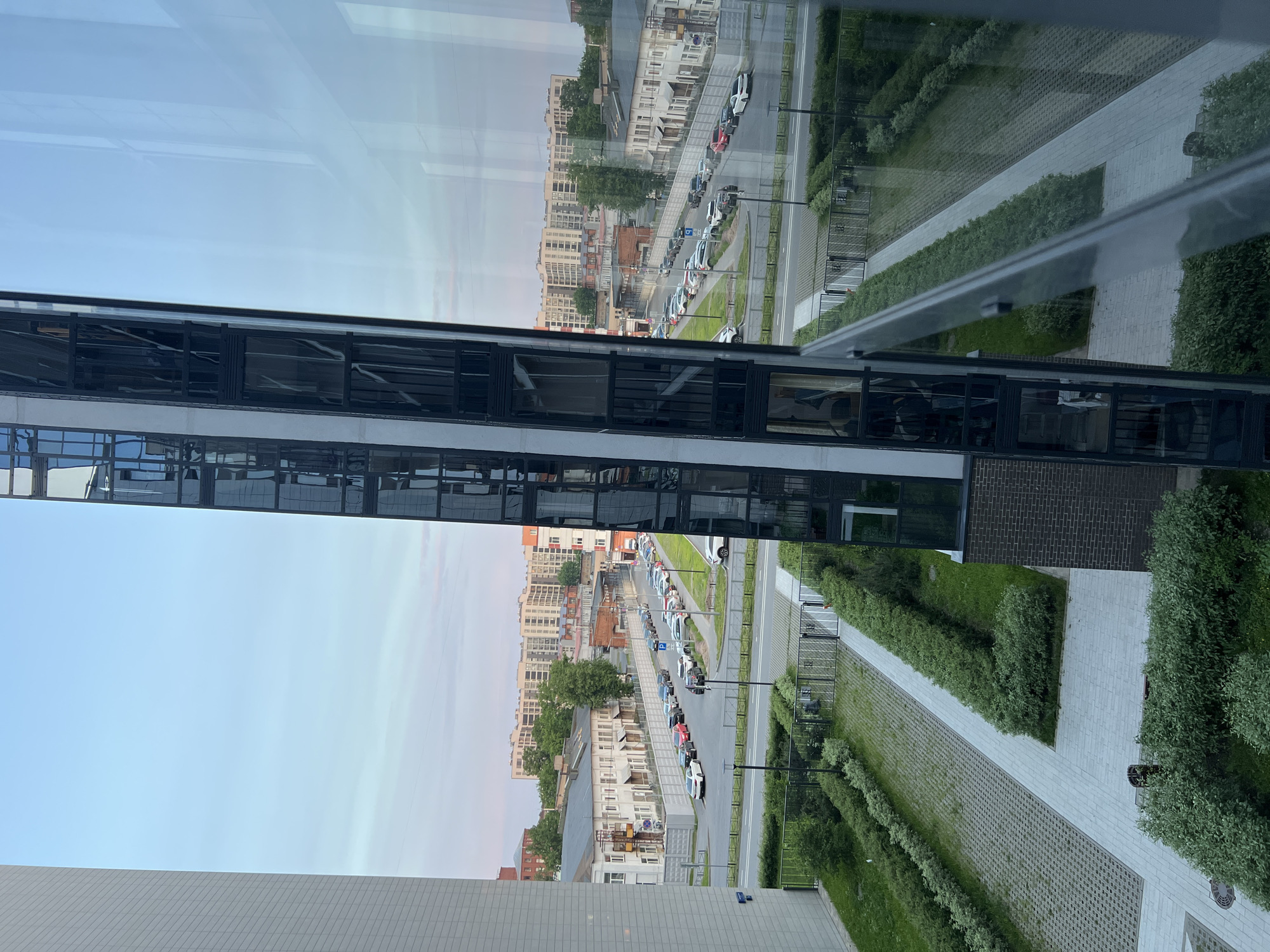 This image captures an elevated view of an urban landscape in Saint Petersburg, Russia, likely from a residential building's window, looking down onto a courtyard and street scene.
In the foreground, a neatly maintained courtyard is visible, featuring a network of light grey paved walkways and areas covered with grey interlocking concrete pavers. Lush green hedges are meticulously trimmed and line the pathways, interspersed with patches of grass. A dark metal fence with gates separates this private green space from the public street beyond. On the far left and right edges, sections of a modern building, from which the photograph is taken, frame the view; the left section is a light-colored, gridded facade, while the right section is dominated by a large, reflective glass surface, showing distorted reflections of the street and sky.
The midground features an asphalt street, curving slightly, with vehicles parked bumper-to-bumper on both sides. The cars are a mix of various models and colors. Along the street, there are paved sidewalks and several streetlights. Behind the parked cars on the left, a row of two-story, light-colored buildings with traditional windows and sloped roofs forms part of an older residential or commercial district. Some of these older structures feature small balconies or external staircases. Further back and to the left, a red brick building is partially visible. On the right side of the street, similar older buildings are present but partially obscured by the window reflection. Mature green trees are scattered throughout this midground, providing foliage.
In the background, beyond the immediate street and older buildings, a cluster of multi-story residential apartment blocks dominates the horizon. These buildings appear to be modern or recently renovated, characterized by their light-colored facades (predominantly beige or off-white) and numerous windows arranged uniformly. The sky above is a pale blue, transitioning to a light pinkish hue near the horizon, suggesting either early morning or late evening. No people are visibly active in the scene. The architecture, combining older Northern European styles with contemporary apartment complexes and urban green spaces, is characteristic of the city of Saint Petersburg.