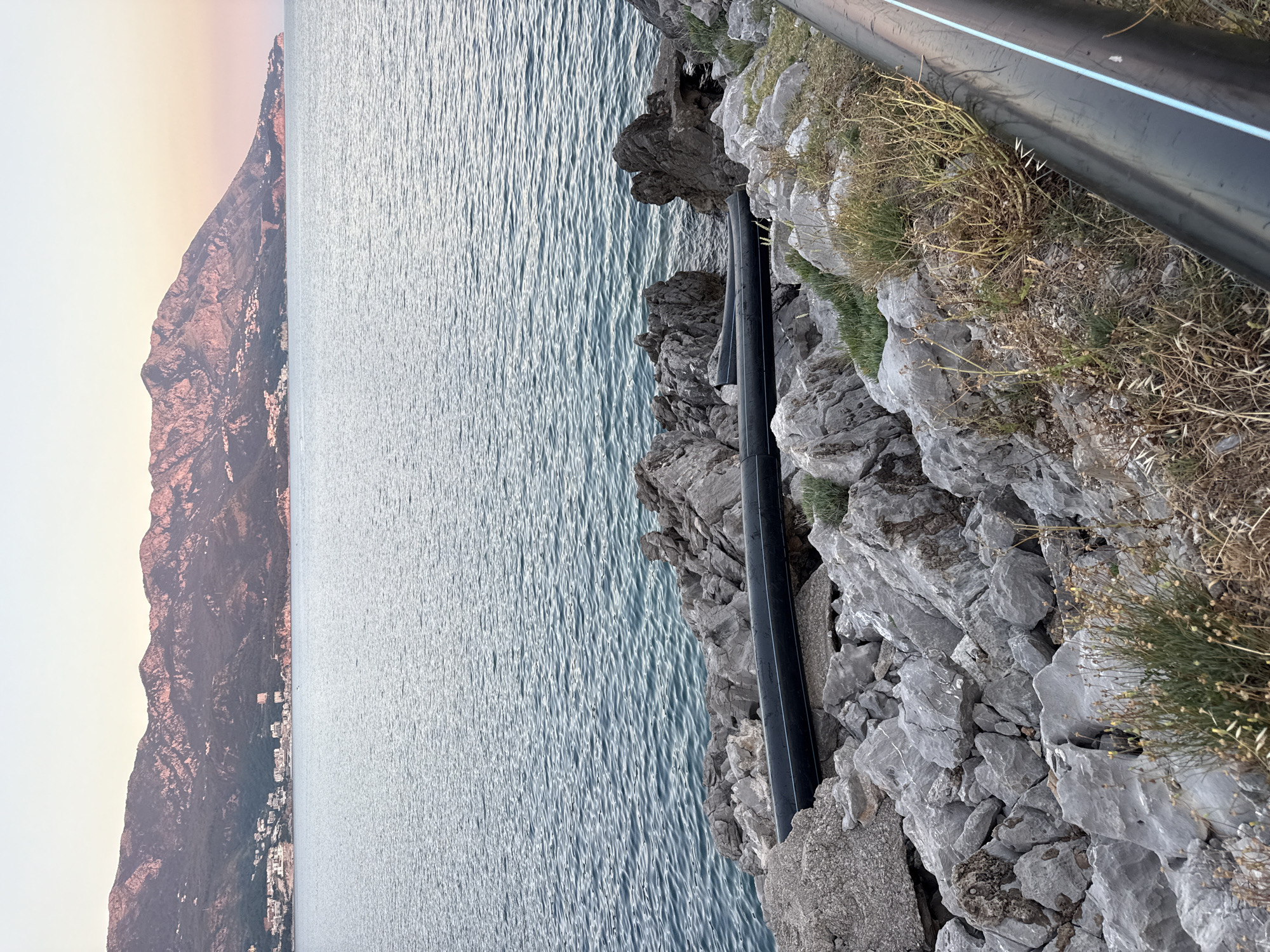 This media file captures a scenic view of the Adriatic Sea and a mountain range in Boreti, Montenegro. The scene is dominated by the vast expanse of the sea, its surface rippling gently under what appears to be soft, natural light. To the left, a majestic mountain rises steeply, its slopes dotted with sparse vegetation and a scattering of buildings, suggesting a coastal town nestled at its base. The sky transitions from a pale, warm hue near the horizon to a lighter tone above, indicating either early morning or late afternoon.
In the foreground, a rocky embankment lines the edge of the water. Prominently displayed on this rocky terrain are two large, black pipes, one running horizontally across the frame and another partially visible beneath it. The pipes are smooth and industrial, contrasting with the rugged, natural texture of the rocks and the dry, wispy grasses growing among them. A thin, light blue stripe runs along the visible pipe, a subtle detail that breaks the monochrome of its surface.
The overall impression is one of a serene coastal landscape, where human infrastructure, represented by the pipes, is integrated into the natural environment. There is no visible activity of people, nor any text present in the image. The time of day is suggested by the soft lighting and the colors in the sky, while the distinct mountain and sea scenery firmly places the location context as Boreti, Montenegro.