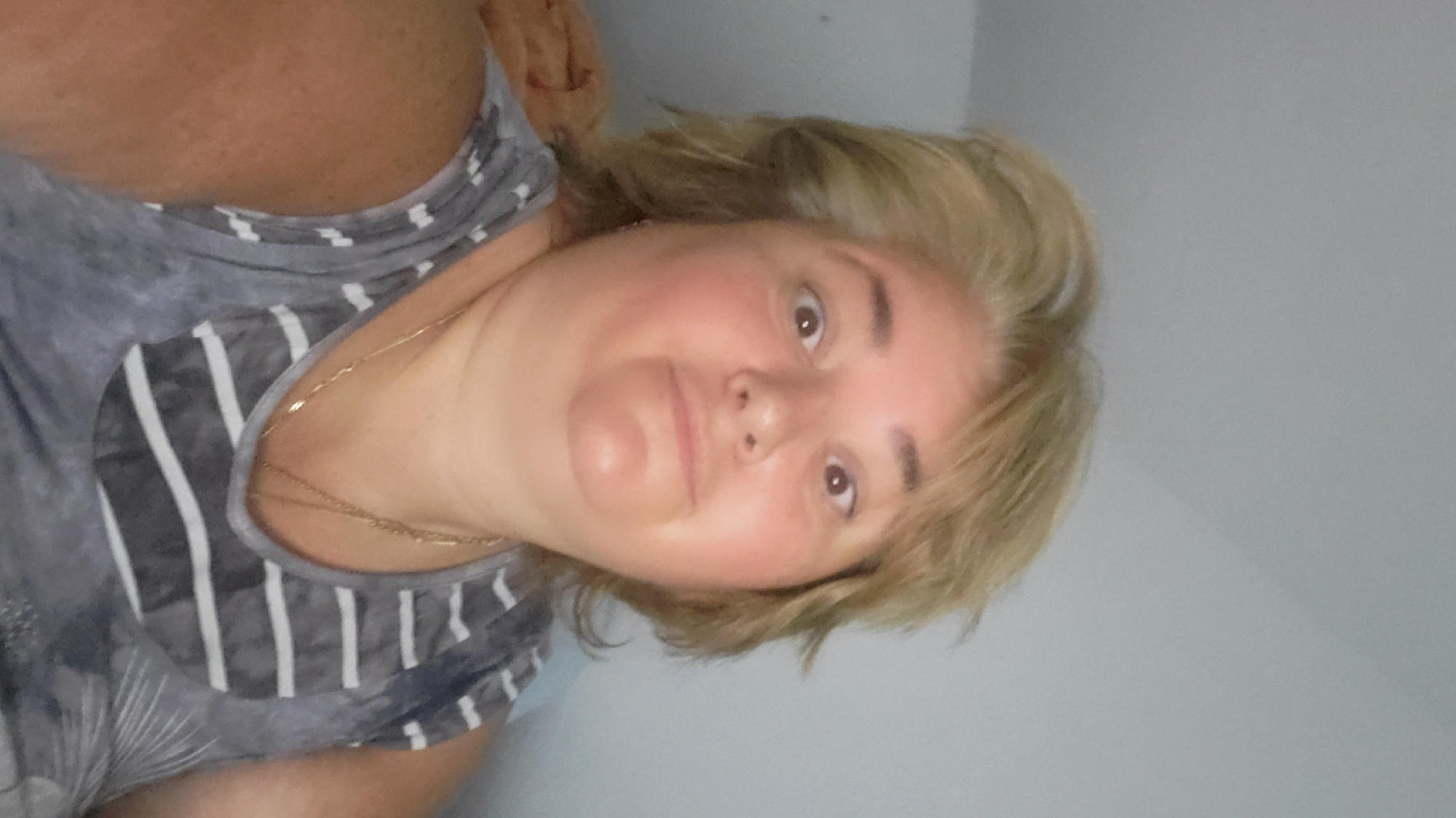 A selfie taken from a low angle shows a fair-skinned woman with blonde hair, fair skin, and light brown eyes. She is wearing a grey and white striped tank top and a gold necklace. The background is a plain grey wall. The image appears to be taken indoors. No specific location in Sasbach, Germany can be determined from the image.