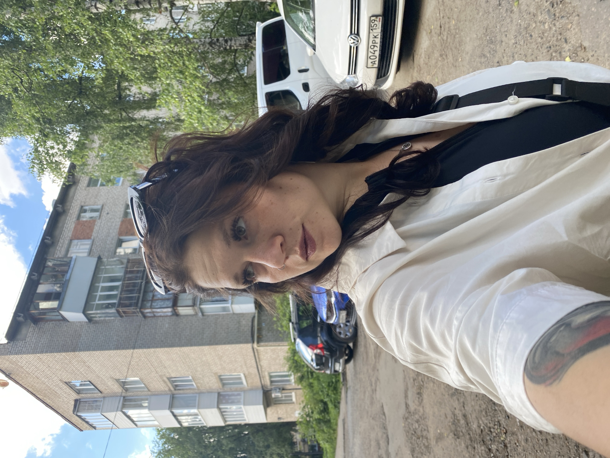A woman is taking a selfie outdoors in Chaikovsky, Russia. She has long, dark, wavy hair and is wearing sunglasses perched on her head. She has on a white collared shirt over a black top and a necklace with a circular pendant. A tattoo is visible on her arm.
In the background, a white van with a license plate reading "A049PK 159" is parked. Behind the van, a blue car is visible. The scene also includes a brick apartment building with several windows and balconies, and lush green trees. The sky is blue with some white clouds. The lighting suggests it is daytime.