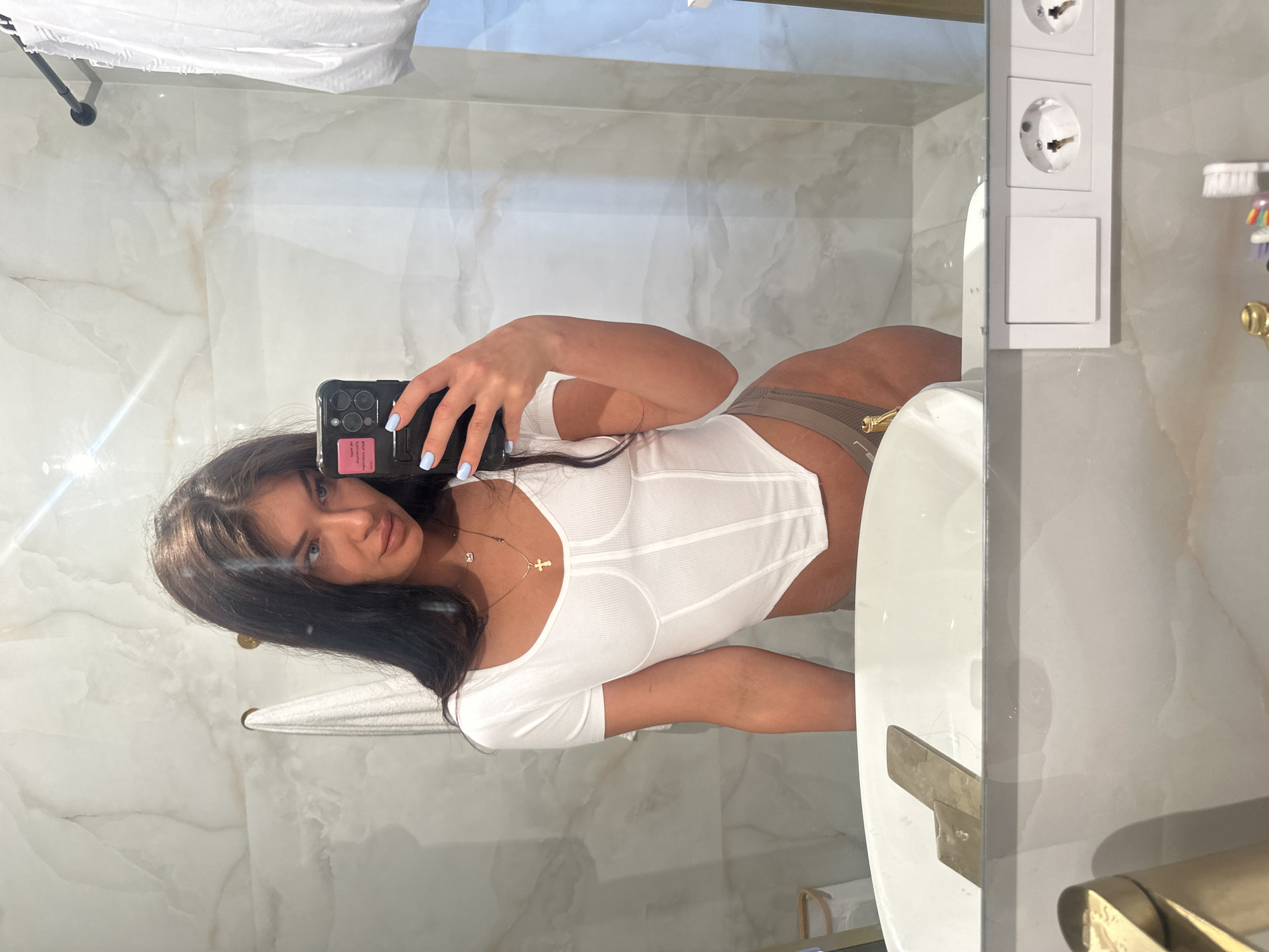 The image, originally rotated 90 degrees clockwise, captures a young woman taking a mirror selfie in a bathroom. She has long, dark hair, blue eyes, and light blue nail polish. She is wearing a white short-sleeved top, possibly a bodysuit or corset-style, and light brown athletic-style bottoms. Around her neck, she wears a gold cross pendant and another small gold pendant.
The woman is holding a dark-colored smartphone, likely an iPhone, in her right hand, which obscures part of her face. A pink sticker is visible on the back of the phone, with partially legible text that includes "EVERYTHING I" and "THE MONEY".
The setting is a modern bathroom featuring polished, light-colored marble-patterned walls. Visible bathroom fixtures include a white sink basin with a shiny gold-toned faucet and handles. A white towel hangs on a rail below the sink. To the right of the woman's reflection, a white toothbrush holder with several toothbrushes of varying colors is visible. Above this, a vertical stack of three white electrical outlets (Type F, or Schuko sockets) is mounted on the wall, indicating a European electrical standard consistent with the given location context of Moscow, Russia. The overall impression is one of a clean, well-lit space, possibly in a hotel or contemporary residence. The woman's expression is neutral as she focuses on taking the photo.