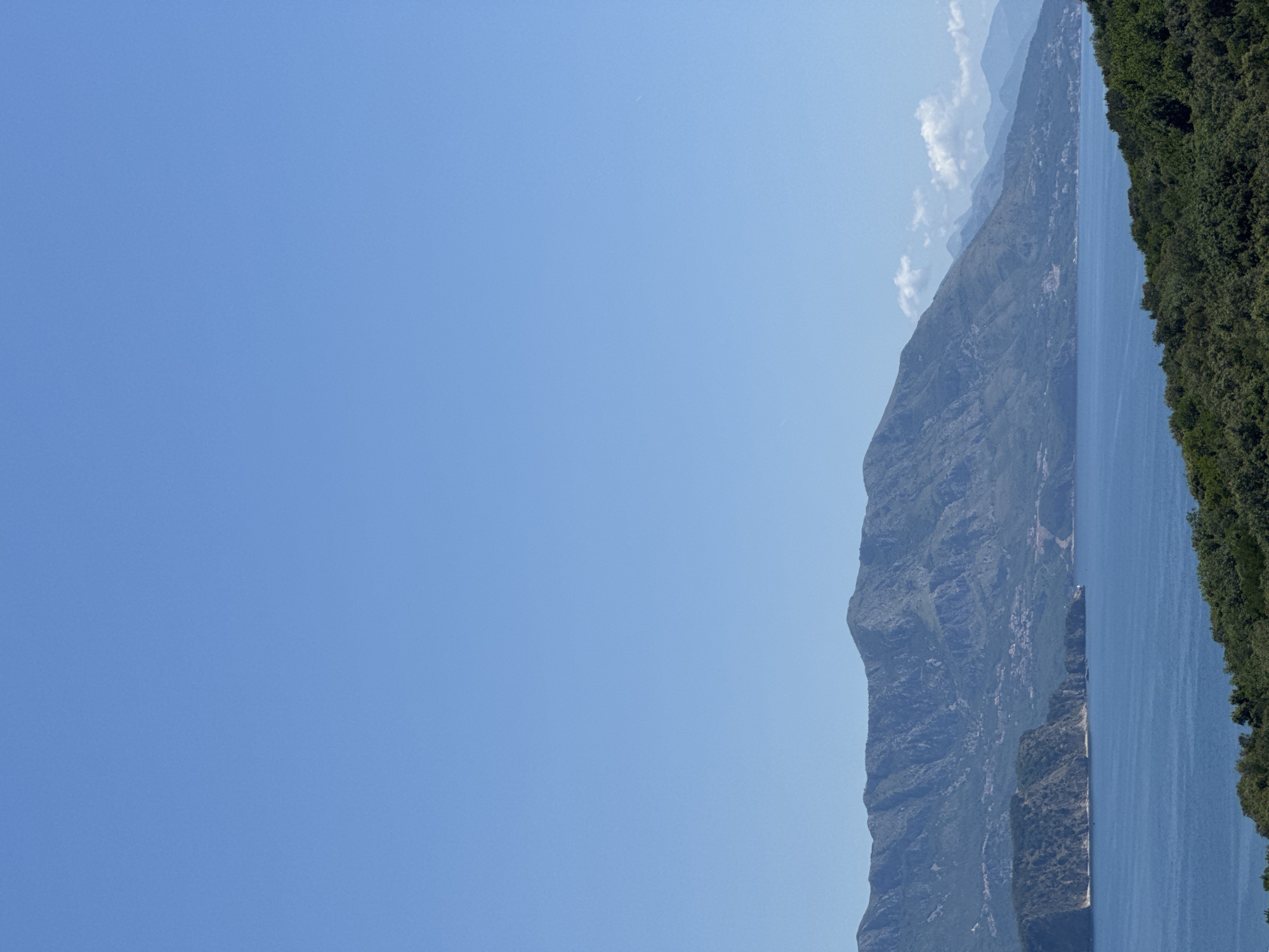 This wide-angle daytime image captures a vast coastal landscape in Kubasi, Montenegro, under a clear azure sky that occupies approximately the top two-thirds of the frame. The horizon line is dominated by a rugged, multi-layered mountain range, appearing dark green to bluish-gray, with settlements visible as small clusters of structures on their lower slopes facing the sea. In the midground, a significant body of tranquil, deep blue water stretches across the scene. A prominent, verdant peninsula or large island with a steep, rocky cliff face extends from the left side into the water. The immediate foreground is densely covered by dark green foliage, suggesting an elevated viewpoint overlooking the coastline. No people, specific objects (beyond natural features and distant buildings), actions, or interactions are discernible within the frame.
