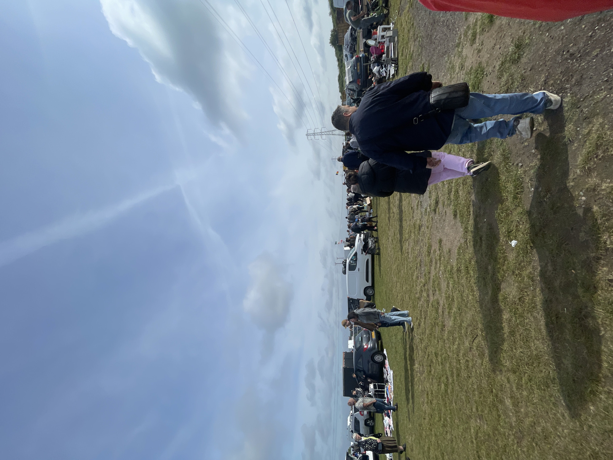 The image depicts an outdoor marketplace in Sevenoaks, United Kingdom, under a cloudy sky. A line of parked vehicles is visible along a grassy field. Numerous individuals are present, some inspecting goods laid out on blankets or tables near the cars. Overhead power lines are also visible. The focus is on the attendees and their activities, set against the backdrop of the field and parked vehicles.