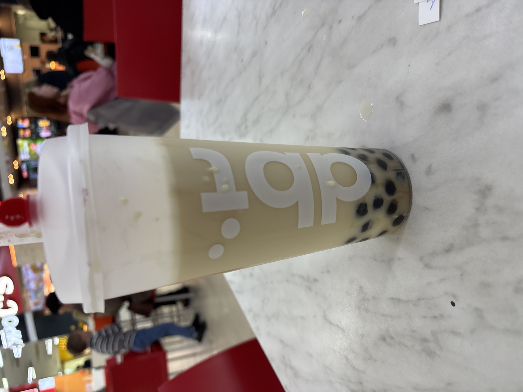 A cup of bubble tea with tapioca pearls is on a marble table. A "daboba" logo is displayed on the cup. A person is seen in the background, along with a seating area and shops. The location appears to be a mall. The city is unknown, and there is no indication of Russia.