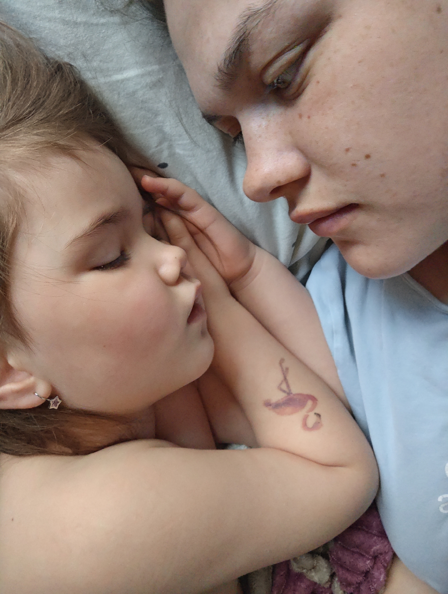 This intimate, close-up portrait captures an adult and a young child lying close together, likely in a bed or on a soft surface, in what appears to be an indoor setting in Chaikovsky, Russia.
The child, a young girl with light brown hair and fair skin, is positioned on the left side of the frame, seen in profile. Her eyes are closed, suggesting she is sleeping or resting peacefully, with her lips slightly parted. She wears a silver star-shaped earring with a purple gem in her left ear. Her right hand is gently placed near her cheek, resting against the adult's arm.
To the right, an adult, likely a woman, gazes down at the sleeping child with a gentle, contemplative expression. She has dark hair, fair skin, and visible freckles on her face. She is wearing a light blue top, and her right hand is tenderly positioned near the child's head. On her left upper arm, a vibrant pink temporary tattoo or decal of a flamingo is visible.
The two individuals are surrounded by soft fabrics, including a grey material resembling a sheet or pillow under their heads, and a textured purple fabric further down. The scene conveys a profound sense of tenderness, peace, and affection between the adult and child, capturing a quiet and intimate moment of rest. No text is visible in the image.