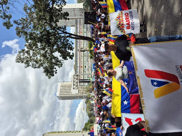 A crowd of people marches, displaying Venezuelan flags and banners. Buildings are visible in the background under a sky with scattered clouds. The banners read "Barrio Nuevo Tricolor" and "Misión Venezuela". The protesters' clothing and the overall ambiance suggest a political demonstration or rally. The perspective is somewhat tilted.