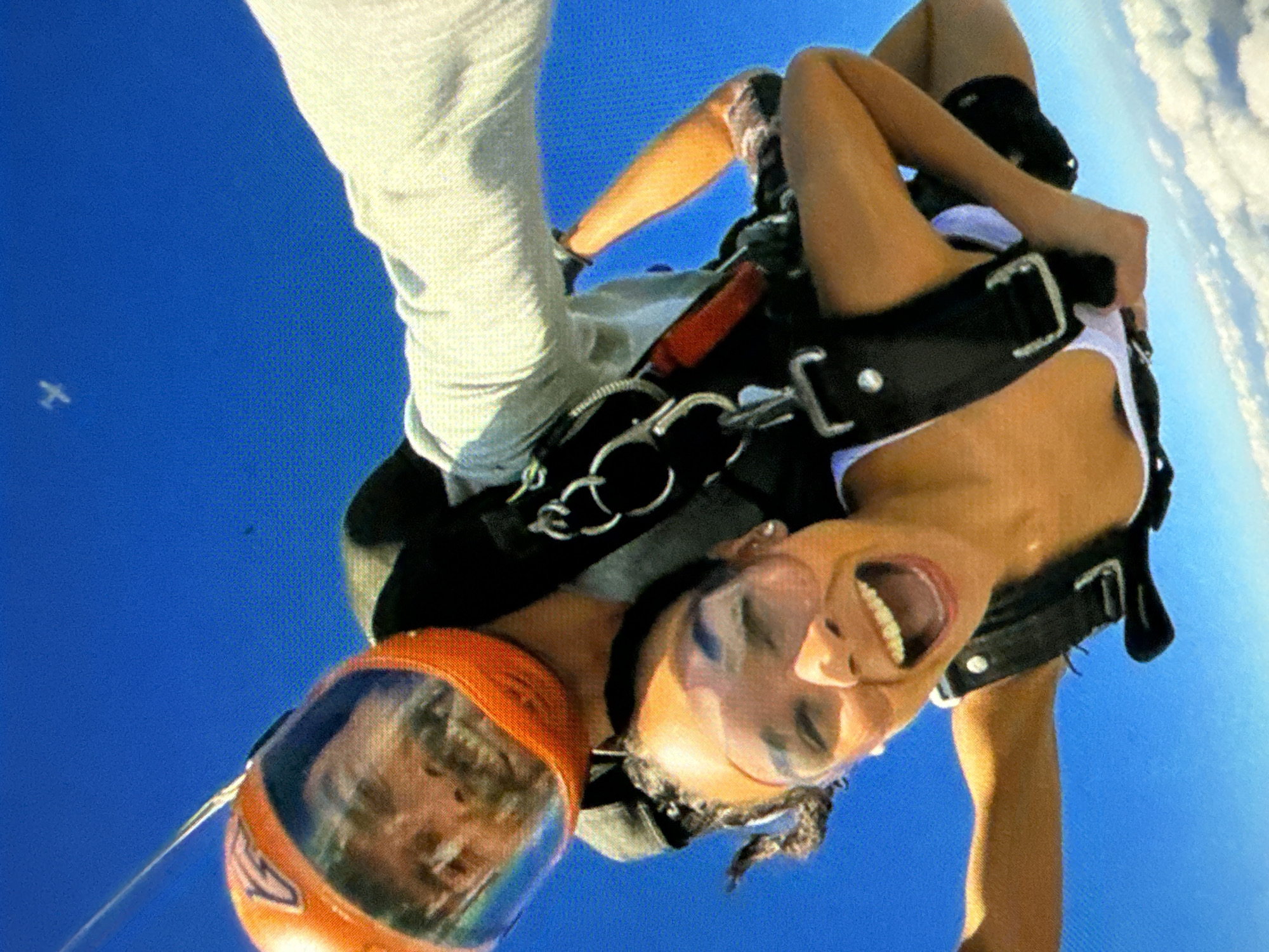 Two individuals are skydiving. The upper person is wearing a white top and a black harness. The lower person is wearing gray pants and an orange helmet. The sky is clear and blue with a few clouds in the distance. A small plane is visible in the upper left corner of the image. The location is Unknown, Unknown.