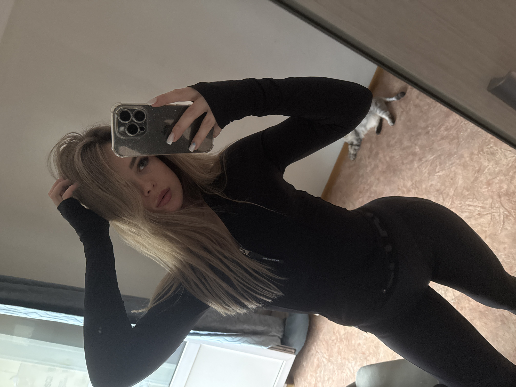 A young woman with blonde hair is taking a mirror selfie. She is wearing a black long-sleeved athletic top and black leggings. Her right hand is raised, touching her hair, and her left hand is holding a smartphone. The background shows a window with curtains, a wall, and a cat lying on the floor.