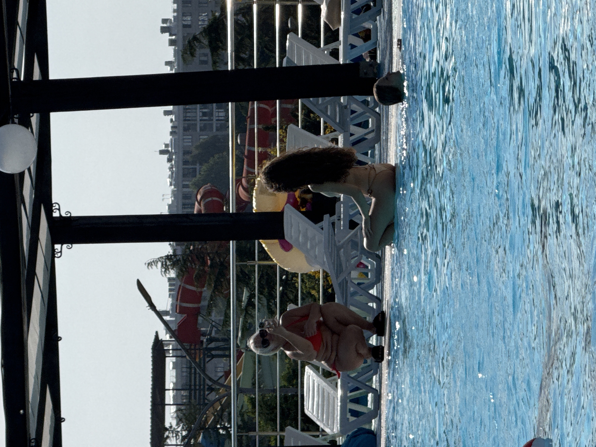 The image depicts a sunny day at an outdoor swimming pool complex, likely in Sevastopol, Ukraine, given the architectural style of the surrounding buildings and the presence of water slides in the background. In the foreground, a bright blue swimming pool occupies most of the frame. On the pool deck, a woman in an orange bikini is crouching and taking a selfie with her phone, wearing sunglasses. Next to her, a younger woman with long dark hair is sitting in a light-colored bikini. Further to the right, a person's head is visible just above the water in the pool. Several white lounge chairs are arranged on the deck. In the background, a section of a water park with red and yellow slides can be seen, along with apartment buildings typical of a city. The scene suggests a recreational environment on a clear day.
