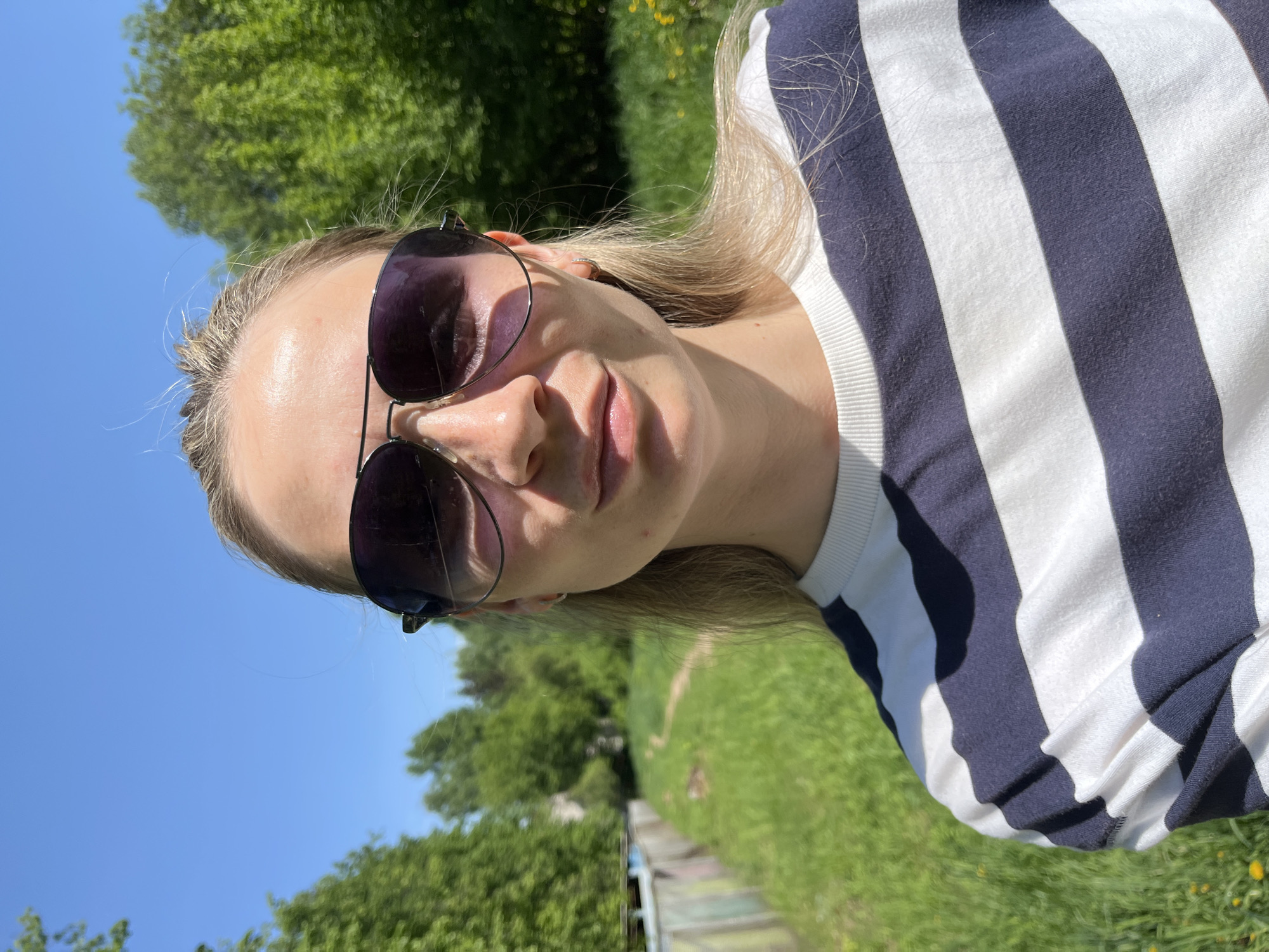 A woman with blond hair is wearing sunglasses and a striped shirt. The background includes green trees, grass, and a blue sky. The location is likely Chaikovsky, Russia.