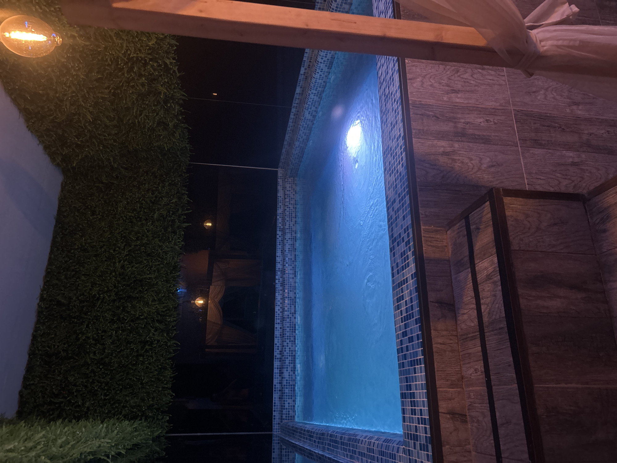 Here is a detailed description of the image:
**Scene:**
The image shows an outdoor pool area at night, likely in a private residence. A rectangular pool with blue tile trim is visible. The pool has illuminated blue water. To the right of the pool, there are wooden steps. The decking area is covered with wood-look tiles.
**Objects:**
- Pool with blue water
- Blue tile trim
- Wooden steps
- Wooden decking
- Faux green wall
- A string of decorative lights, and a single visible bulb.
- Wooden beam.
- Sheer fabric canopy.
**Environment:**
The scene is illuminated by decorative lights and the underwater pool lighting. The background is dark, suggesting it is night.
**Location:**
Without further information, it's not possible to pinpoint the exact location in Sofia, Bulgaria. The image depicts a private residence, which could be in many residential areas of the city.