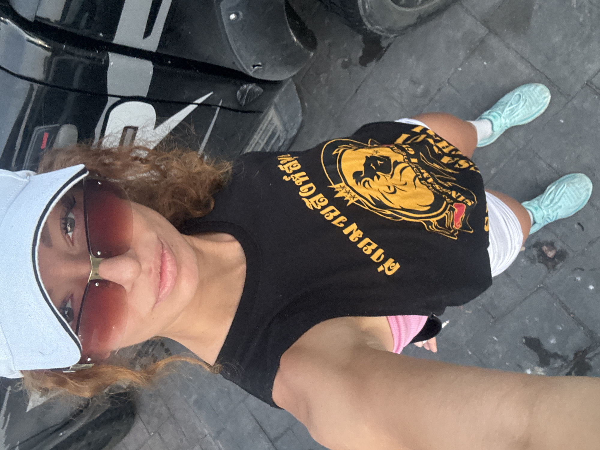 A woman is taking a selfie on a brick sidewalk in what is likely Baan Chaweng, Thailand. She is wearing a light blue baseball cap, sunglasses, a black t-shirt with gold graphics, white shorts, and light blue sneakers. Part of a black vehicle with a white Nike swoosh is visible next to her.