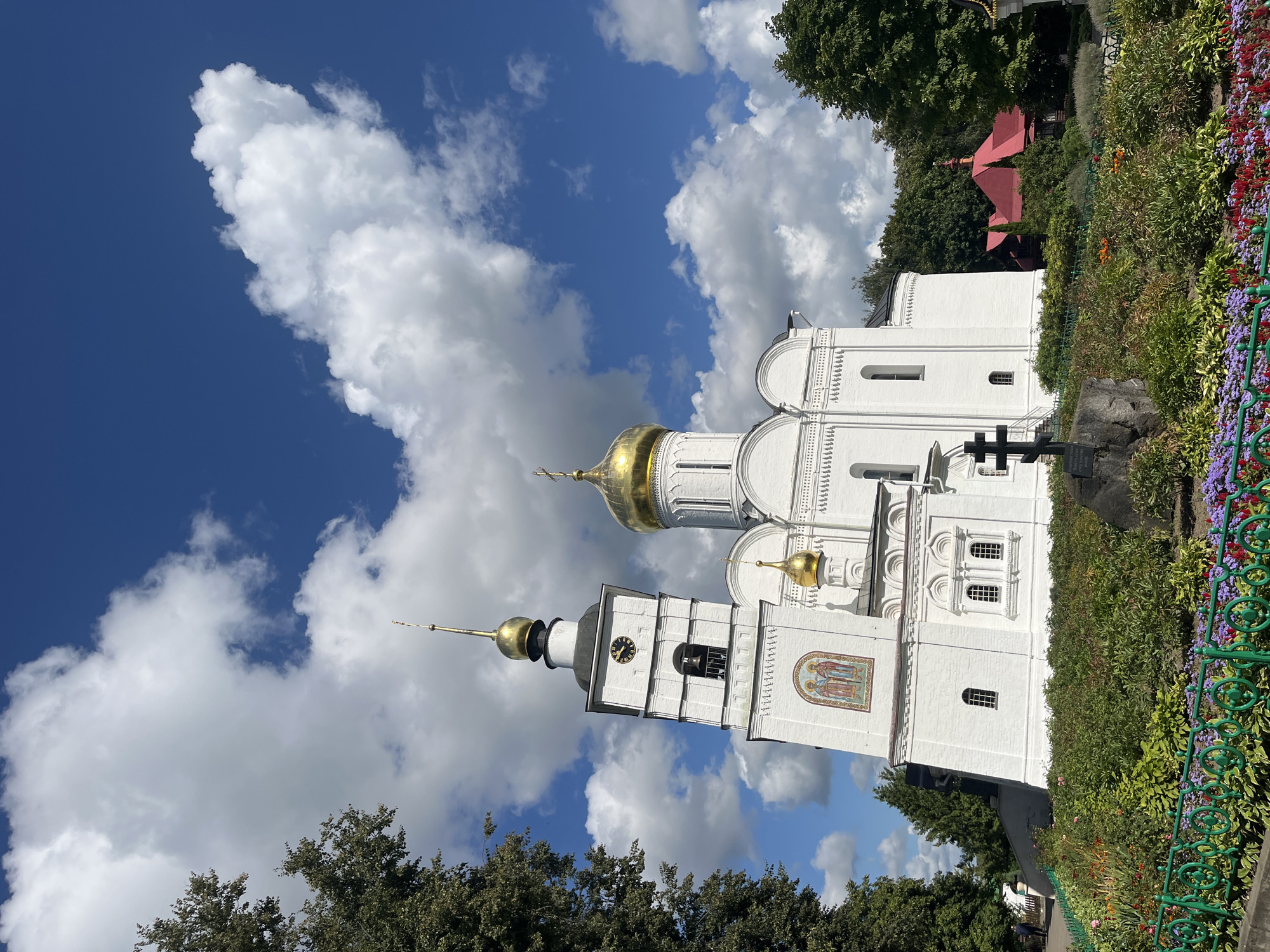 A bright, clear day with abundant white clouds in a deep blue sky provides the backdrop for the Assumption Cathedral and its bell tower in Dmitrov, Russia. The white cathedral, adorned with a golden dome and a smaller golden cupola, is a striking sight, as is the tall, white bell tower. The bell tower features a clock face and a prominent bell within its open belfry. Below the bell tower, a mosaic depicting two figures is visible. In the foreground, a lush garden with a variety of colorful flowers and dense green foliage is bordered by a decorative green fence. A black cross stands prominently on a rock in front of the garden. To the right of the cathedral, a building with a red roof can be seen nestled amongst the trees. A large tree with dark green foliage is on the left side of the image, and other trees with similar foliage are visible behind the cathedral and the red-roofed building. The scene is calm and serene, capturing the architectural beauty of the religious site and its surrounding natural environment.