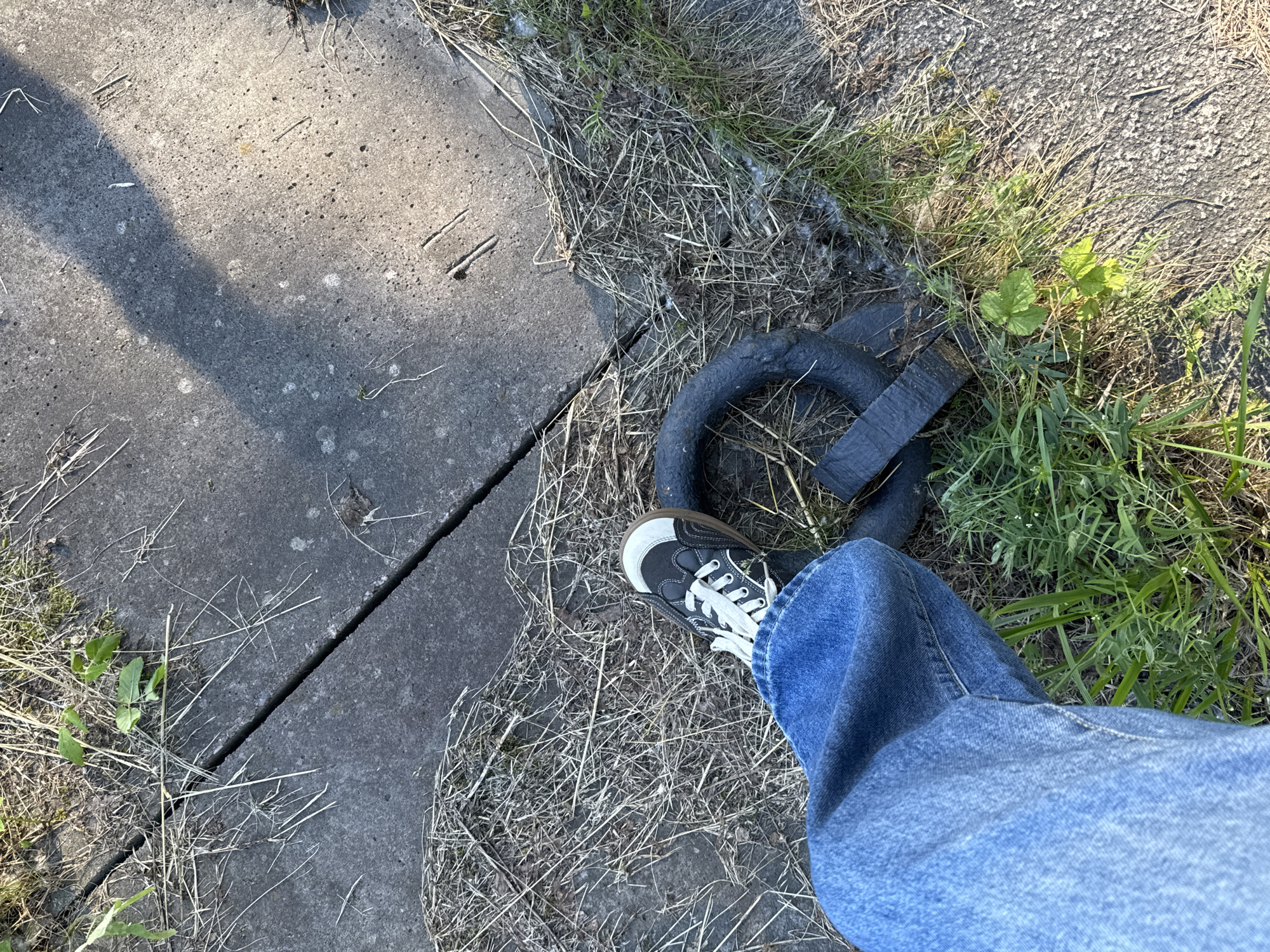 A person's left leg, clad in blue denim jeans and a black and white sneaker, is positioned in the lower left quadrant of the frame, stepping on a dark, circular object with a ridged surface. The object appears to be a piece of heavy machinery or industrial equipment. The ground is composed of cracked concrete slabs and dry, sparse vegetation, suggesting an outdoor urban environment. A shadow, possibly cast by a nearby structure or object, stretches diagonally across the concrete. The scene is lit by natural sunlight. The specific location in Saint Petersburg, Russia, cannot be determined from the visual information provided.