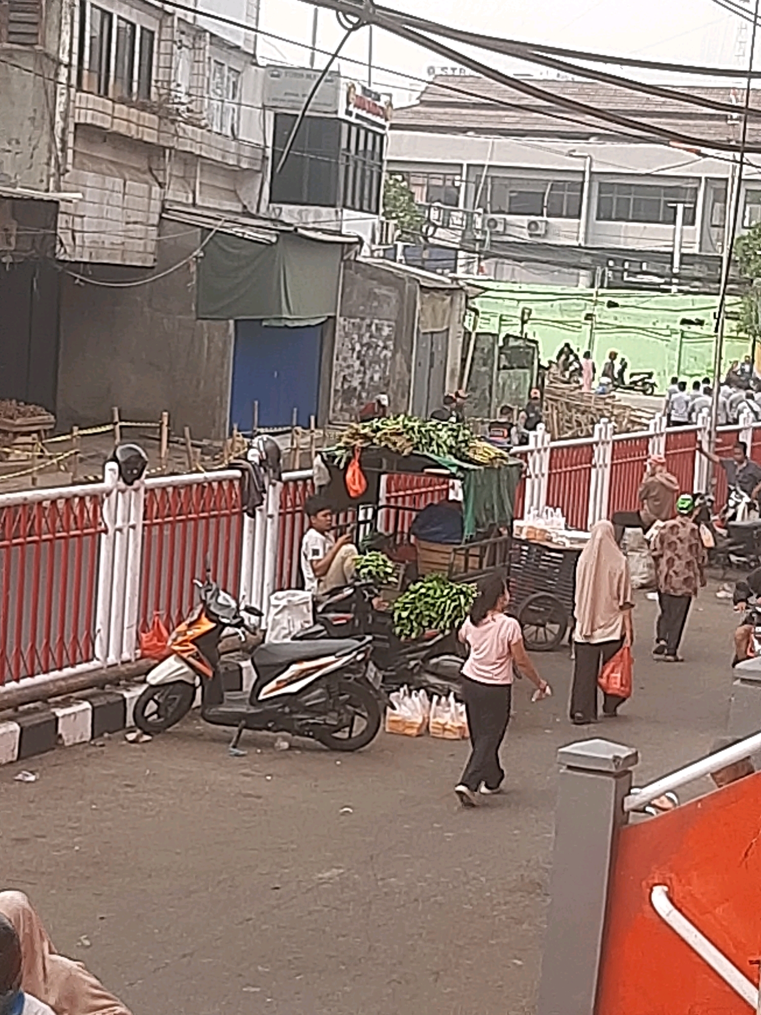 The image depicts a street scene in Bekasi, Indonesia, likely a marketplace or roadside area with several people and vehicles. In the foreground, a person in a pink shirt and black pants walks away from the camera, passing by a scooter. To the left, another scooter is parked. Behind them, a street vendor's cart is visible, laden with produce, and a person sits near the cart. A red and white fence runs horizontally across the mid-ground. Beyond the fence, several more people are visible, some walking and some standing. Buildings, appearing to be a mix of commercial and residential structures, form the backdrop, with power lines crisscrossing overhead. The overall impression is one of a bustling urban environment.