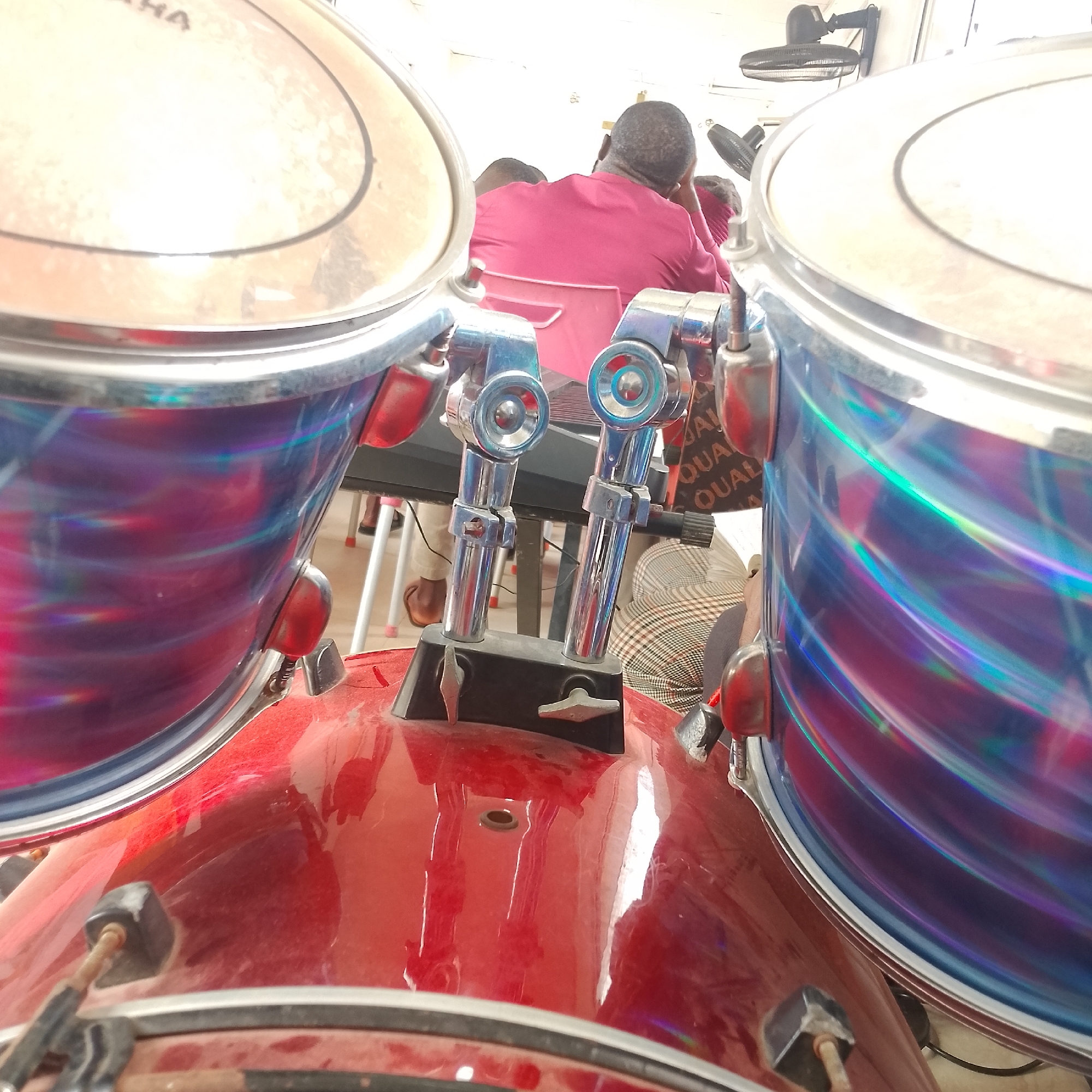 In Iranla, Nigeria, a close-up perspective of a drum set is presented. The image is dominated by a red bass drum in the foreground, its reflective surface showing streaks of light. To the left and right, portions of two tom-tom drums are visible. These drums have a metallic sheen with an iridescent wrap that displays swirling patterns of purple, blue, and green. The drum hardware, including lugs and mounting brackets, are metallic and silver-toned. In the background, several individuals are seated. One person in a vibrant pink or maroon outfit is the most prominent, seen from behind, with their head resting on their hand. Other figures are partially obscured by the drums, but their presence suggests a gathering or performance. A black wall-mounted fan is visible on the ceiling in the upper right corner, and a keyboard or electronic instrument is partially visible behind the tom-tom drums. The overall environment appears to be an indoor space, possibly a church or community hall, suggested by the lighting and the presence of multiple people.