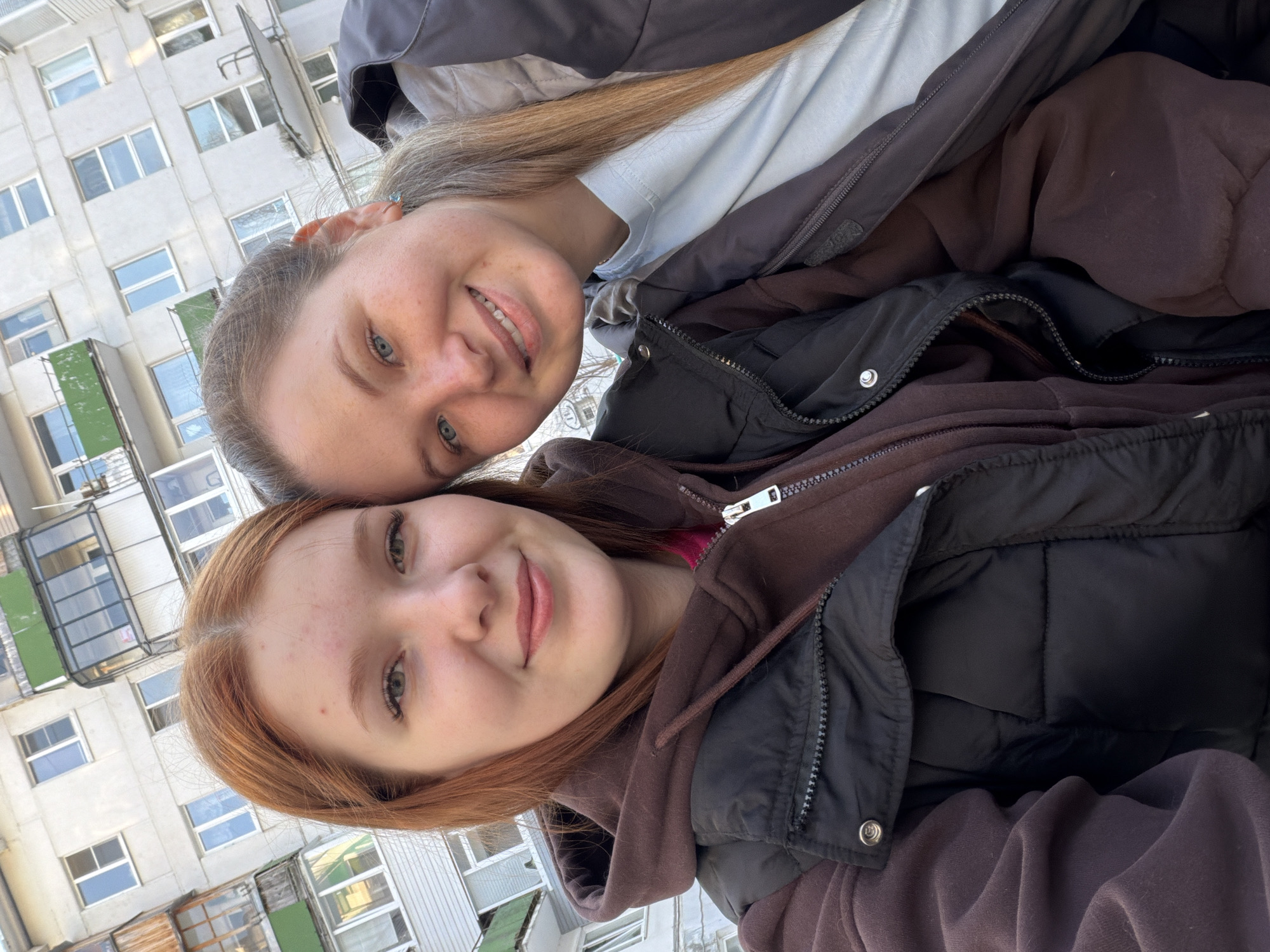 Two young women are posing for a selfie in front of a multi-story residential building. The building has numerous windows and what appear to be balconies. The woman on top has blonde hair, and the woman on the bottom has red hair. They both are wearing layered clothing appropriate for cool weather, including jackets and hoodies. The setting appears to be a residential area in a city. The location is Chaikovsky, Russia.