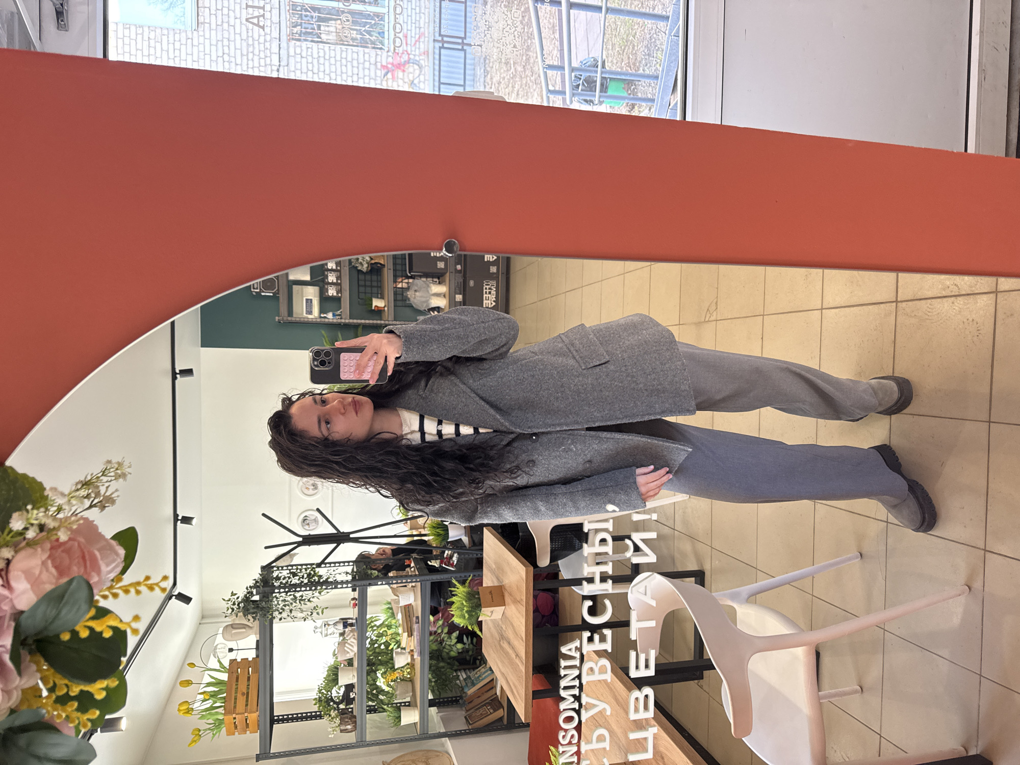 A young adult female is depicted taking a mirror selfie inside "INSOMNIA Coffee & Bloom" located at Vokzalnaya Street, 46A, Chaikovsky, Perm Krai, Russia. She has long, dark, wavy hair and is wearing a grey double-breasted blazer over a striped sweater, grey trousers, and dark grey chunky-soled boots. Her right hand holds a black smartphone encased in a pink pop-it style cover.
The interior reflected in the large, arched mirror with a reddish-orange frame reveals a brightly lit commercial space. Features include a multi-tiered black metal shelving unit displaying numerous potted plants, decorative items, and various merchandise. A wooden table with black legs and white chairs is visible in the background. A wall sign behind the table reads "INSOMNIA" and "С ВЕСНЫ, ЦВЕТАЙ!" (meaning "FROM SPRING, BLOOM!"). The ceiling is white with black track lighting fixtures, and the floor is composed of light-colored tiles. A section of a dark green accent wall is visible.
Through a window reflection on the right, part of the building's exterior and street scene can be observed. This includes a light-colored brick building with barred windows, a grey metal railing, and a green trash receptacle on what appears to be a paved or ground area.