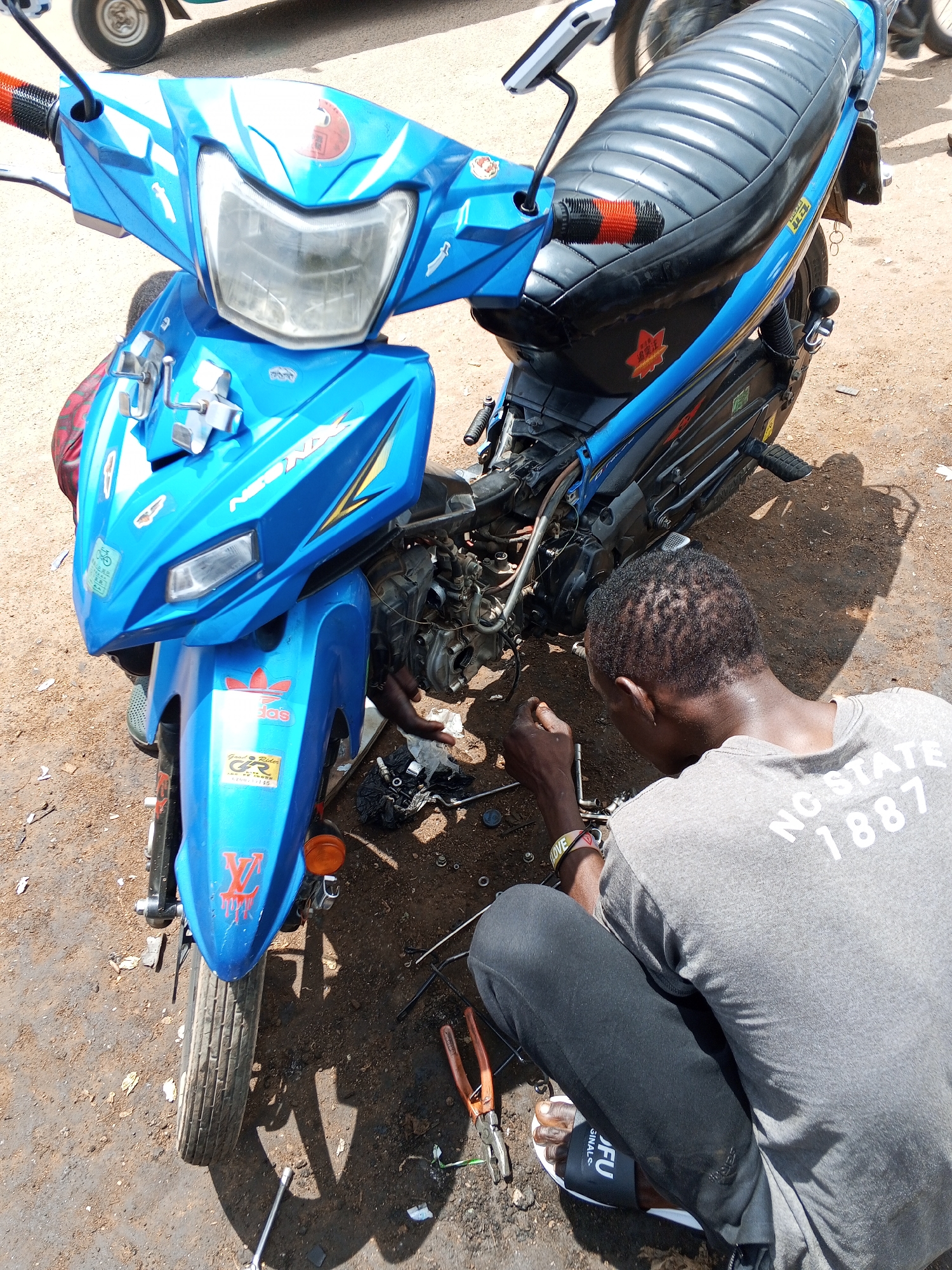 An overhead medium shot captures a man working on a blue scooter in Jos, Nigeria. The man, wearing a gray t-shirt with "NO STATE 1887" printed on the back and black pants, kneels on the ground, his back to the camera. He is using tools, including pliers and a wrench, to repair the scooter. The scooter is partially disassembled, with its engine exposed. The scene is set on a dirt ground, with various tools and parts scattered around. The sunlight casts a long shadow of the scooter on the ground.