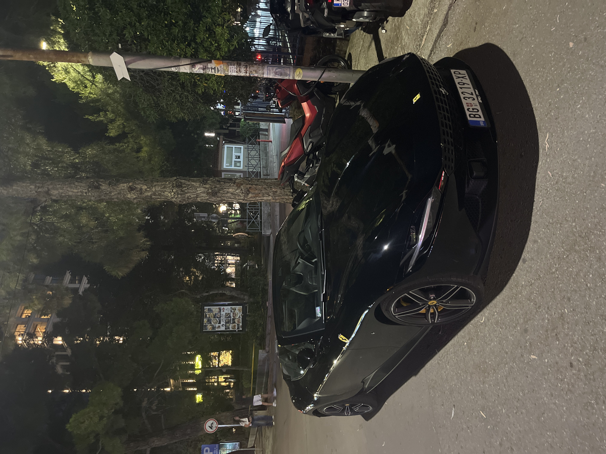 In the city of Unknown, Unknown, at night, a black convertible Ferrari Roma is parked on a paved street. The car has a yellow Ferrari badge on the hood and a license plate that reads "BG 3219 XP". The interior of the car is visible, with black seats. To the right of the Ferrari, a red motorcycle is parked, and further to the right, a black scooter is parked. In the background, there are trees and a building with illuminated windows. Two people are standing on the sidewalk to the left of the car, one wearing a white shirt and black shorts, and the other wearing a white t-shirt and jeans. A street light and several signs are also visible.