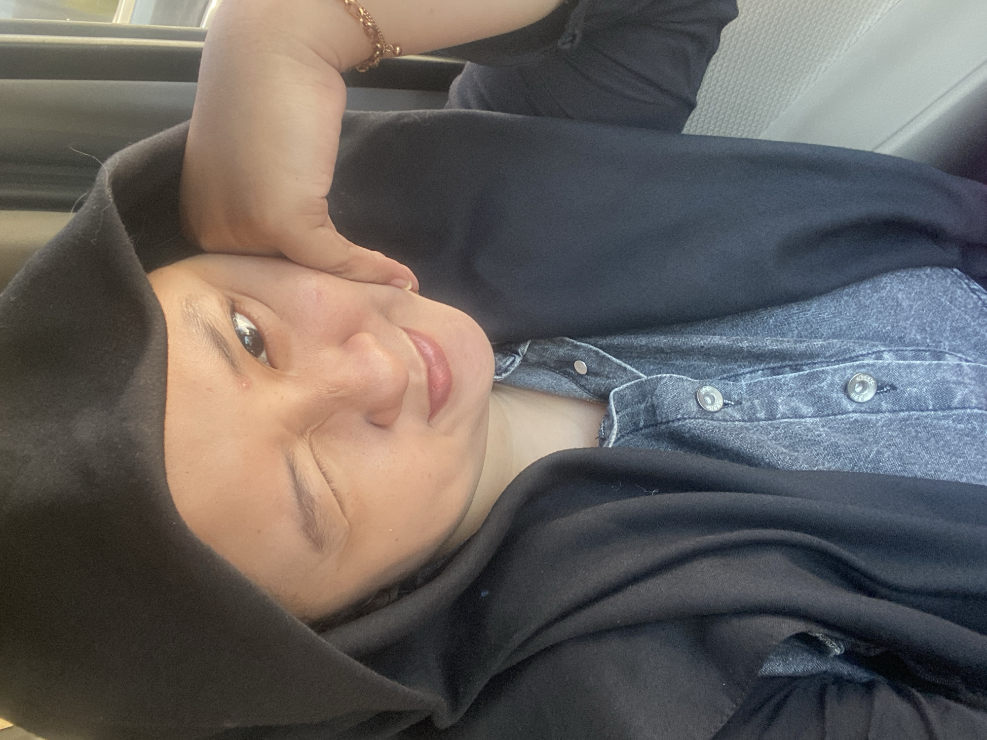 The image is a close-up selfie of a person wearing a black hooded garment and a denim shirt. The person has their right hand resting on their cheek, and their left eye is winked shut. They are looking towards the camera with their right eye open. The background appears to be the interior of a vehicle, with a blurred window and curtain visible on the right side. The person is wearing a rose gold bracelet on their left wrist. The overall lighting suggests it is daytime. There is no specific information about the location in Ngrame, Indonesia.