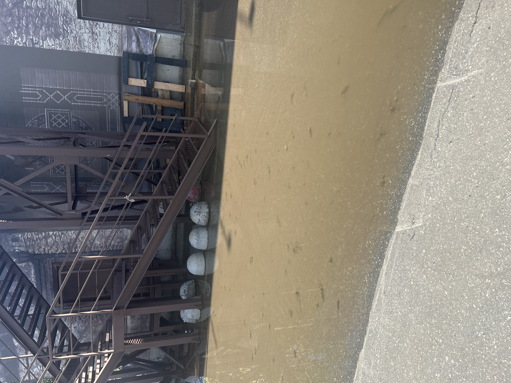 This image depicts a flooded area, likely an alleyway or courtyard, adjacent to a brick building with a metal fire escape. The water, which appears murky and brown, covers the ground, reflecting the surrounding structures. Several rounded concrete bollards are partially submerged in the water near the base of the fire escape. In the background, the building's facade is visible, constructed of brick with some decorative elements. Part of the metal structure of the fire escape, with its grated steps, is prominent in the foreground. The ground outside the flooded area is paved with asphalt. The scene is illuminated by natural daylight. No people are visible in the image. The location is identified as a part of the city of Perm, Russia.