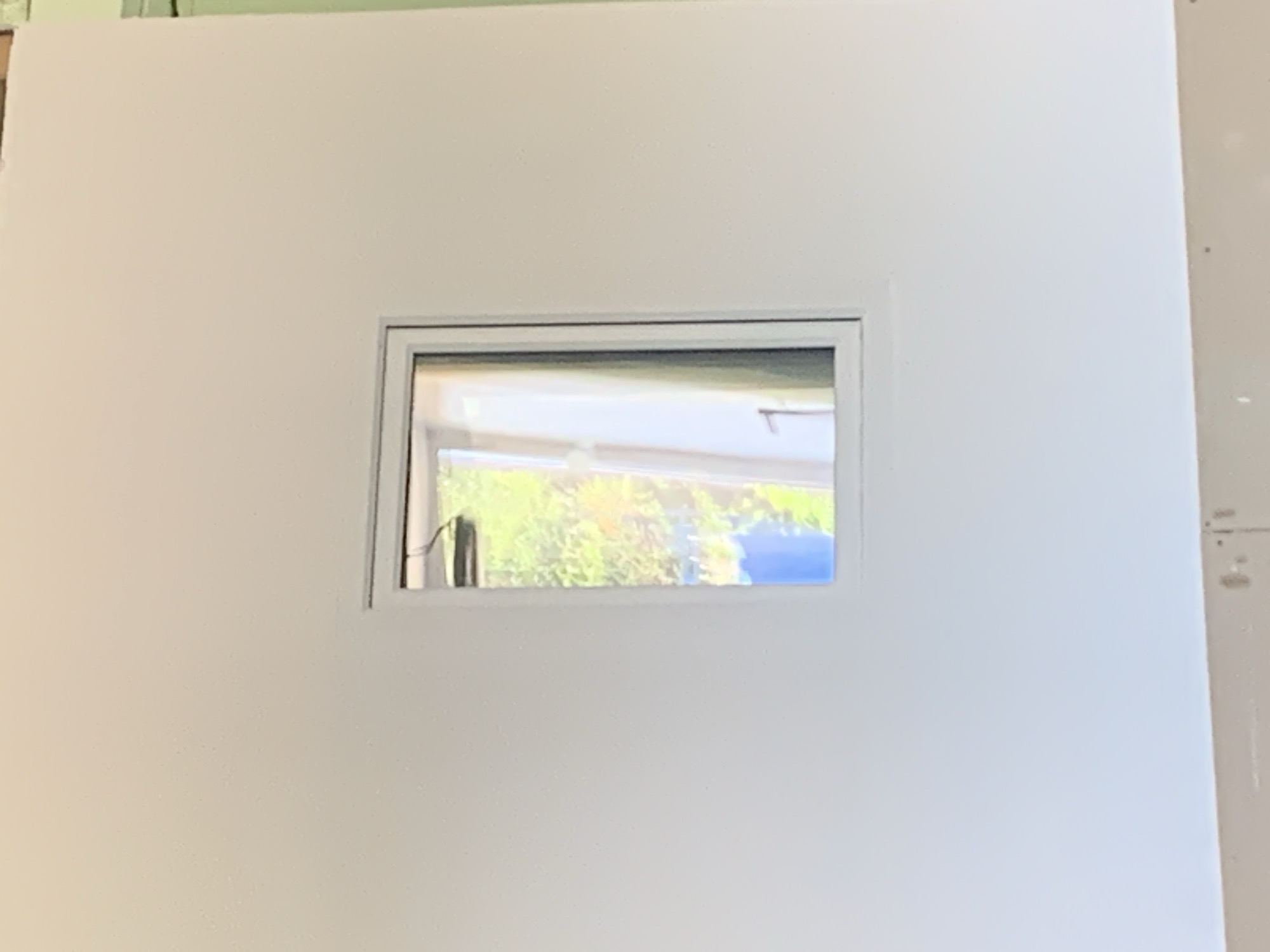 The image shows a white wall with a small, horizontal window in the center. The window frame is light gray. Through the window, a sunlit scene with green foliage and a blue sky is visible. The foliage appears to be trees or bushes. The wall itself is smooth and appears to be drywall. The right edge of the wall shows a slightly different texture and color, possibly indicating the edge of the wall or a transition to another surface. There are small, dark dots visible on this edge, which might be fasteners or imperfections. The lighting suggests it is daytime. No people, text, or specific activities are depicted. The setting appears to be indoors, looking out onto an outdoor scene.