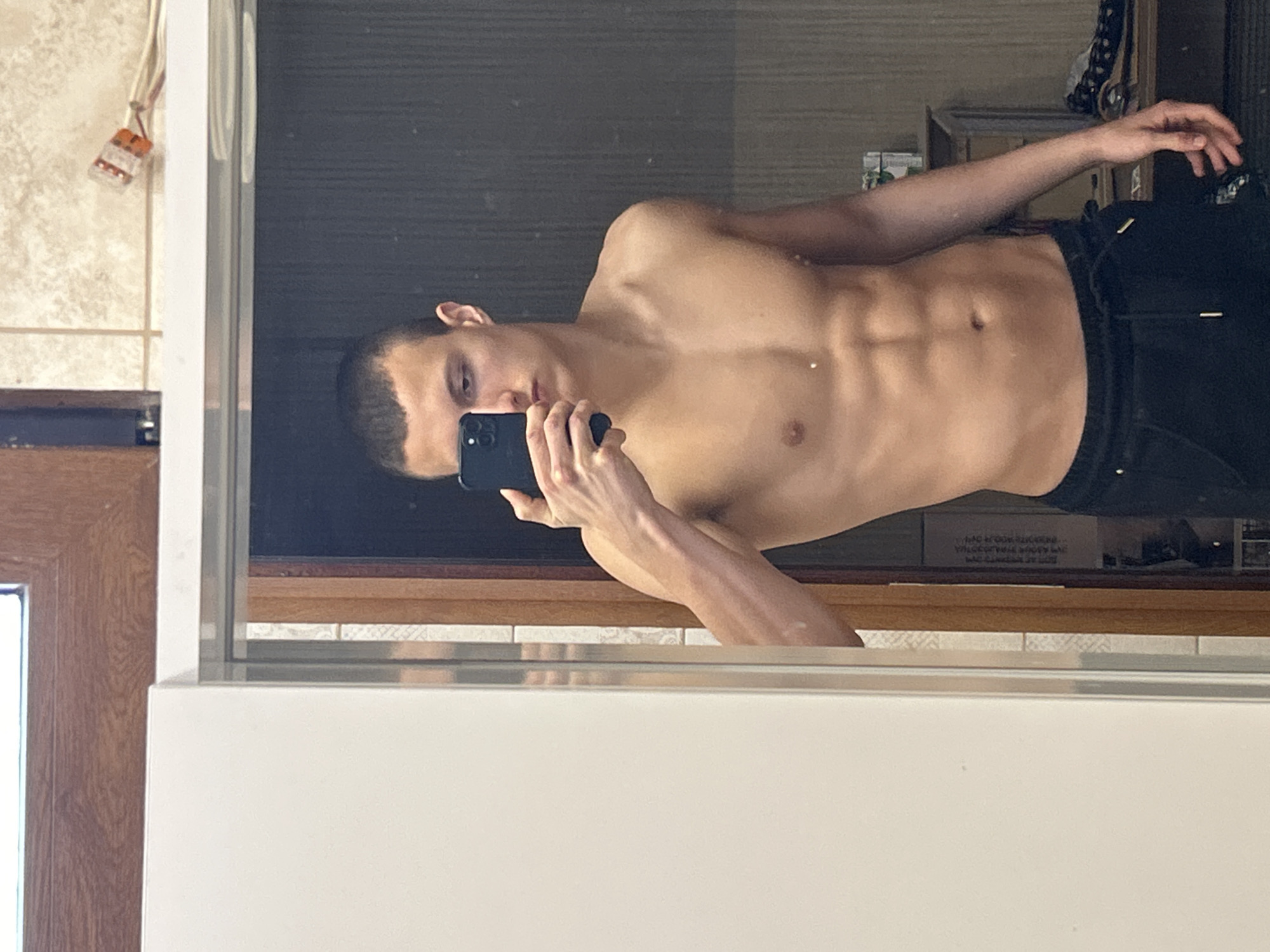 A shirtless young man is taking a mirror selfie. He's fair-skinned with dark hair and is wearing dark pants. He's holding a dark-colored smartphone. The setting appears to be a bathroom; there are tiles and wood trim visible, along with electrical components near the upper-left corner. The location in the city of Burgas, Bulgaria is not discernible from the image.