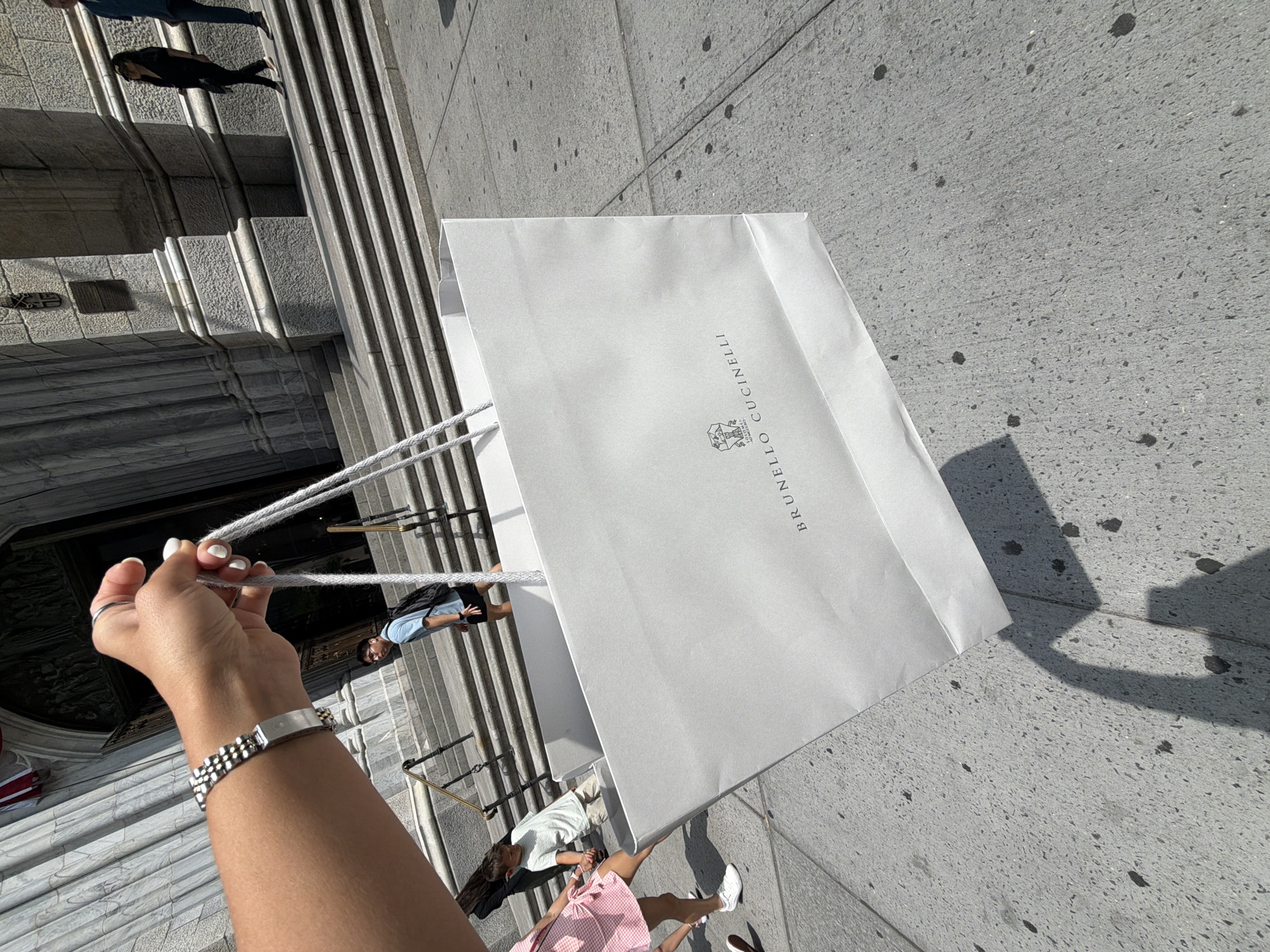 A person's hand, adorned with a silver link bracelet and two rings, holds the handles of a large white shopping bag. The bag displays the "BRUNELLO CUCINELLI" logo. In the background, a grand stone building with large columns and a wide set of stairs is visible. Several people are walking on the stairs and sidewalk in front of the building, including a young boy in a striped shirt and shorts, a person in a blue shirt and black shorts with a backpack, and two people in dark clothing. The ground is paved with grey concrete. The scene appears to be a sunny day, indicated by the strong shadows cast by the building and the people. The architectural style of the building suggests a location in a major city with historic structures, possibly in New York City, United States of America.