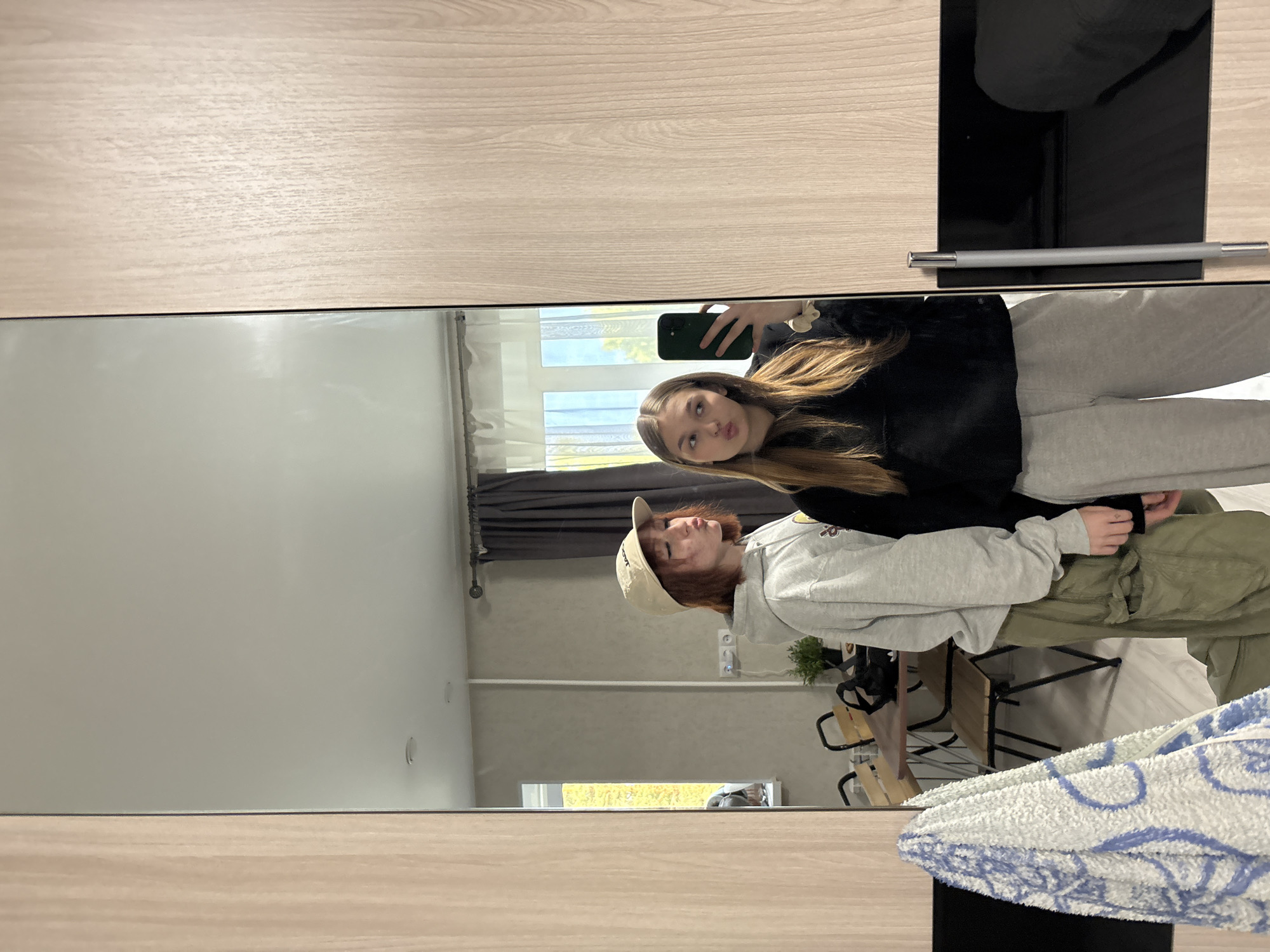 The image is a mirror selfie taken indoors, likely in a bedroom or living area. Two young women are reflected in a full-length mirror. The woman on the left has short, reddish-brown hair and is wearing a light grey hooded sweatshirt, olive green cargo pants, and a beige baseball cap. She is puckering her lips and looking towards the mirror. The woman on the right has long, blonde hair and is wearing a black hooded sweatshirt and light grey sweatpants. She is holding a dark green smartphone in her right hand and is also puckering her lips, looking at the phone screen. In the background, reflected in the mirror, is a room with light grey walls, a window with dark grey curtains, a dining table with chairs, a potted plant, and an electrical outlet. To the left of the mirror, a blue and white patterned towel is hanging. The area outside the window appears to be a street or residential area with trees and buildings.
This specific location is not determinable from the image alone. Therefore, it is impossible to definitively place this scene in Chaikovsky, Russia, or any other city.