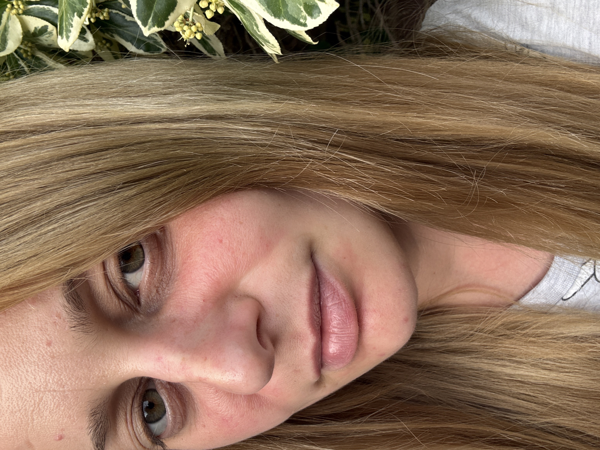 A close-up, angled portrait of a woman with fair skin and blonde hair. The woman's face is the primary subject, featuring light green eyes, a slightly closed mouth, and a pink undertone to her complexion. Her hair, long and straight, partially frames her face. In the upper left corner, foliage with green and white variegated leaves is visible. A small section of what appears to be a grey or light-colored shirt is visible on the right side. The image does not depict any identifiable features that would pinpoint a specific location within Hazlemere, United Kingdom. There is no indication of any actions or interactions. The context and details provide a representation of the woman's appearance.