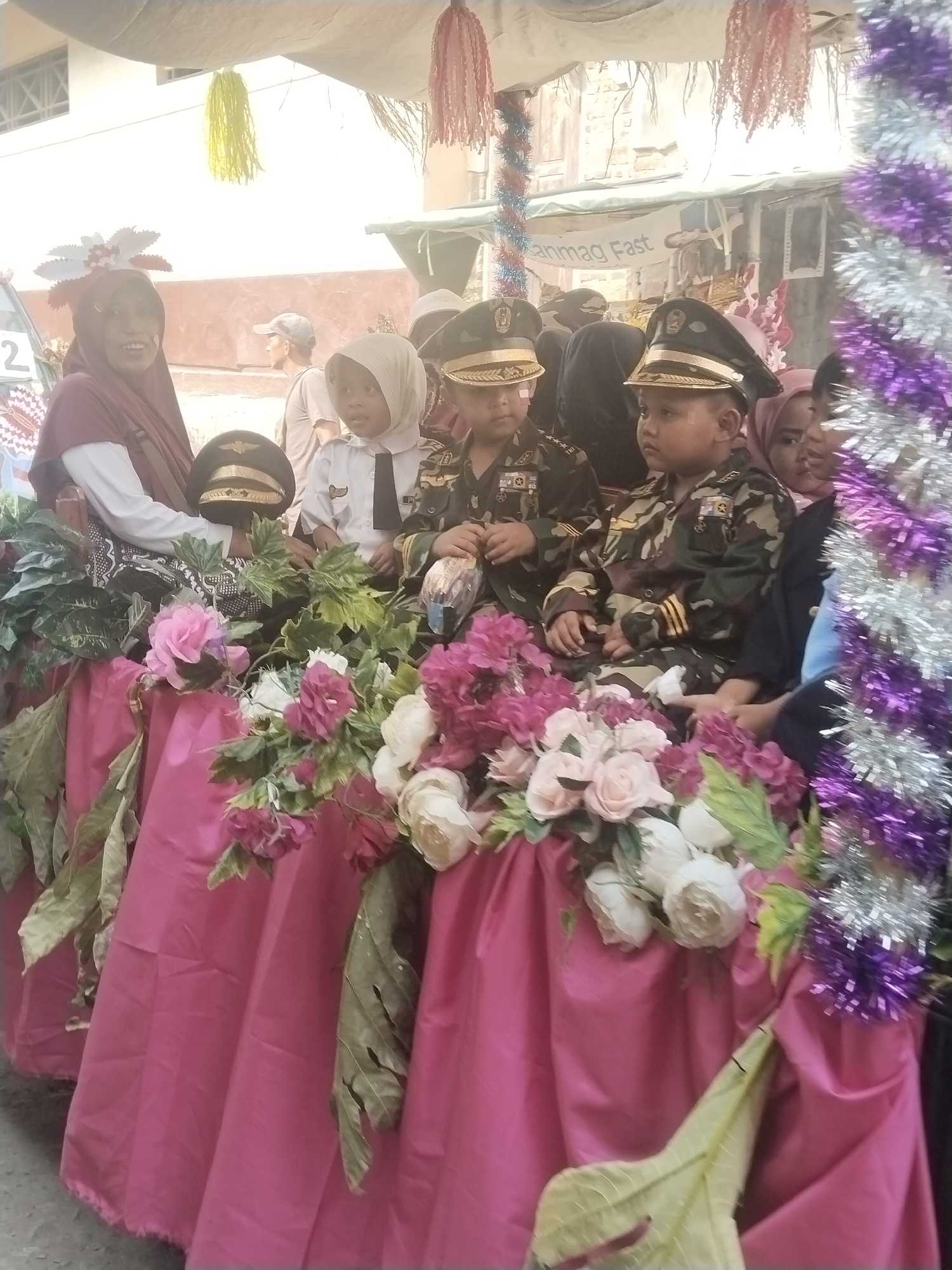 In Jembayat, Indonesia, a group of children dressed in military-style uniforms and a woman in a hijab are seated on a decorated float. The float is draped in pink fabric and adorned with artificial flowers and greenery, as well as purple and silver tinsel. Two boys in camouflage uniforms with caps are prominently featured, along with a younger child in a white shirt and tie, also wearing a cap. The woman in the hijab is seated to the left of the children, wearing a red head covering and a white top. In the background, a banner reads "anmag Fast," and other individuals are partially visible. The overall scene suggests a festive event or parade.