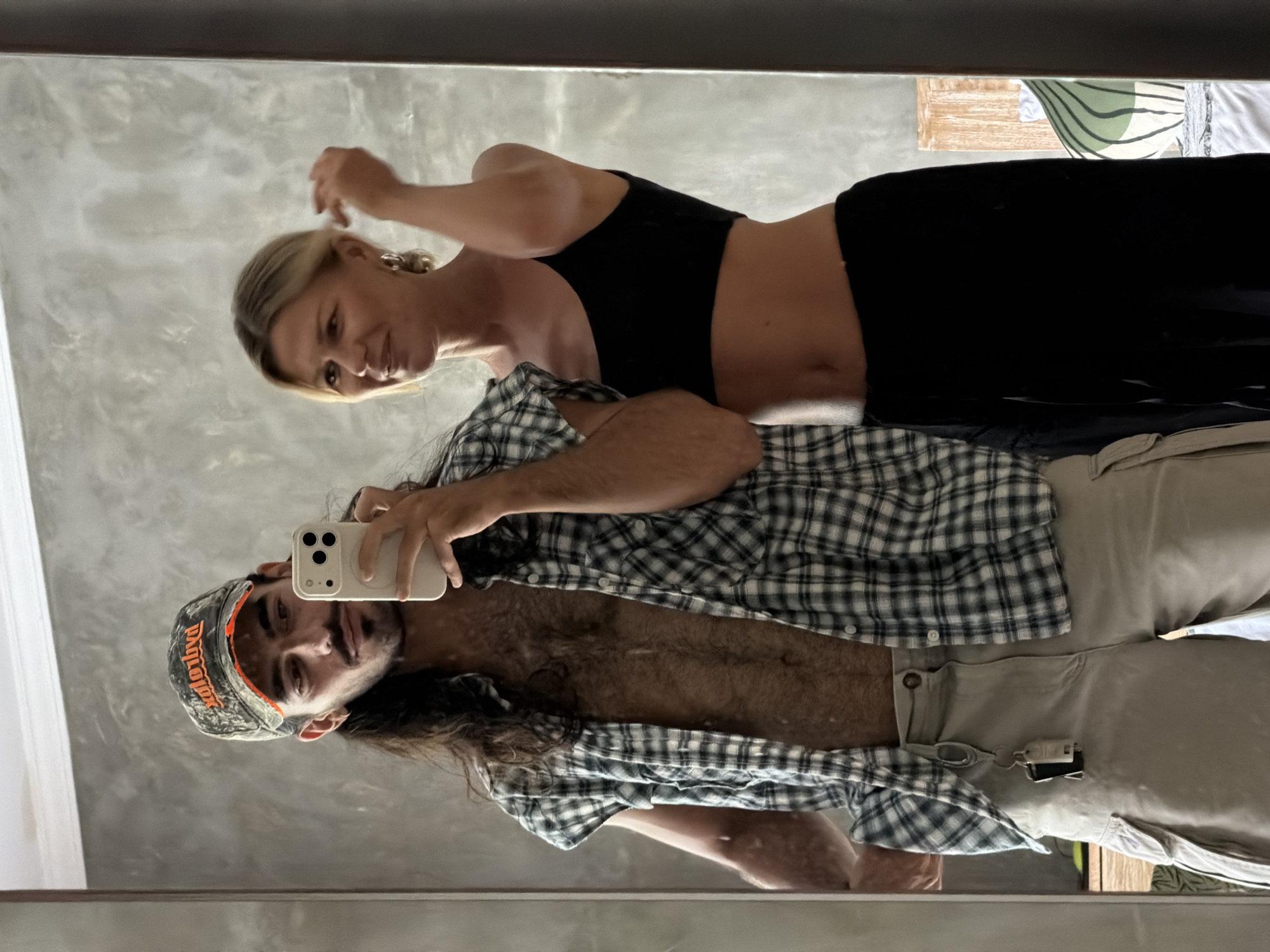 Two individuals, a man and a woman, are captured in a mirror selfie inside what appears to be a modern indoor setting, possibly in North Kuta, Indonesia. The overall scene is casual and relaxed, with a neutral-toned, possibly textured wall as the background reflected in the mirror.
The woman is positioned behind and slightly to the side of the man, looking towards the camera with a subtle smile or smirk. She has blonde hair pulled back and is wearing a black crop top and black trousers. Her right arm is slightly raised, suggesting she might be adjusting her hair or making a casual gesture.
The man, holding a light-colored smartphone with multiple cameras (likely an iPhone) to take the mirror selfie, stands in front. He has long, dark hair, a beard, and a mustache. He is wearing a camouflage-patterned baseball cap with orange text, partially visible as "Ducks Unlimited." He wears an open black and white plaid shirt, revealing his bare, hairy chest, and light-colored khaki or beige trousers. A small, light-colored object, possibly a keychain or lighter, is clipped to his trousers near his hip. He maintains a neutral to slightly smiling expression.
The activity is a relaxed mirror selfie taken by the man, with the woman casually posing alongside. The general lighting suggests it is daytime. In the upper right corner of the reflection, a bed or furniture with a green and white striped pattern is partially visible. Both subjects convey a comfortable and content mood.