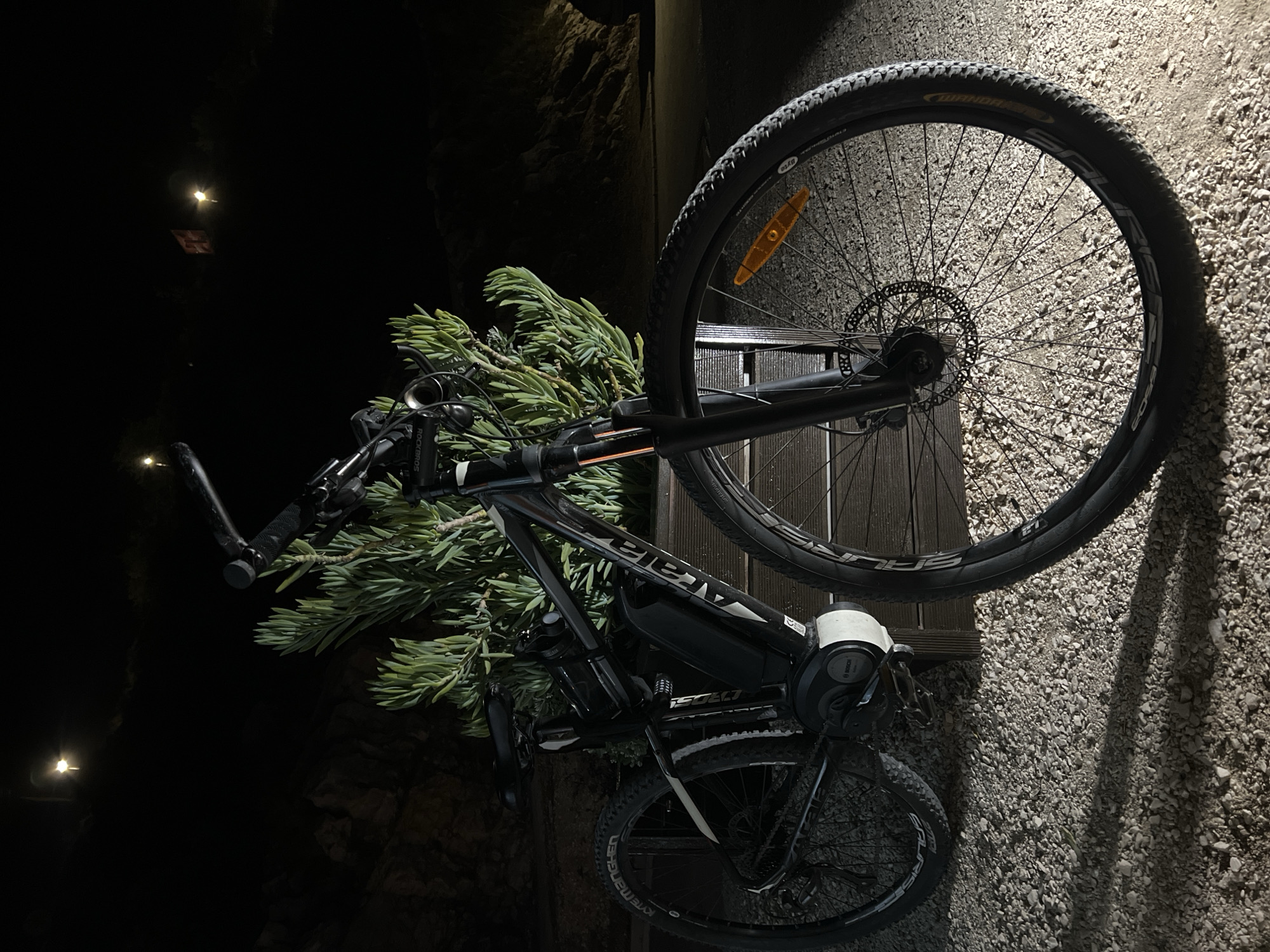 An electric mountain bike with green foliage strapped to the handlebars leans against a wall in Boreti, Montenegro. The bike is black and white with the brand "ISOLT" visible on the frame. A white cylindrical object is mounted near the seat post. The front wheel is suspended in the air, angled towards the top right of the image. The rear wheel is on the ground, resting on a surface that transitions from gravel to planks of wood. The wall behind the bike is textured, possibly concrete or stone. In the background, to the left, is a dark, shadowed area with faint lighting.
