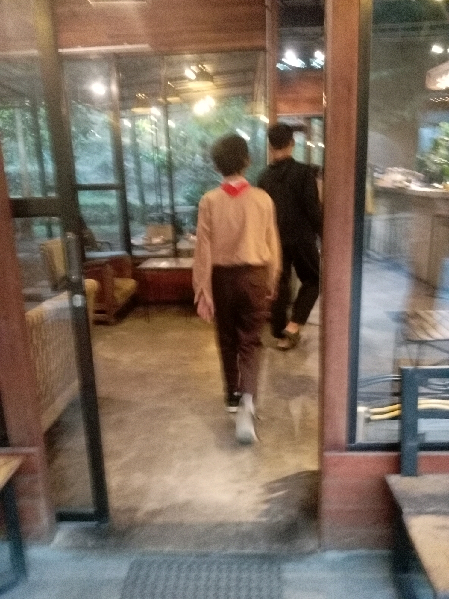 The image depicts two individuals walking away from the camera within an indoor setting that appears to be a cafe or restaurant. The person in the foreground is wearing a light brown long-sleeved shirt and dark brown pants, with a red scarf or bandana around their neck. They are walking towards the right side of the frame. Behind them, the second person is wearing a black hooded jacket and dark pants. They are walking in the same direction. The environment features large glass windows with a view of greenery outside, wooden architectural elements, and concrete flooring. Furniture, including chairs and tables, is visible in the background through the windows. The lighting suggests it is either evening or an overcast day. The scene is blurry, indicating motion or a low-resolution image. The provided context indicates this location is in Krisik, Indonesia, though this is not visually discernible from the image itself.