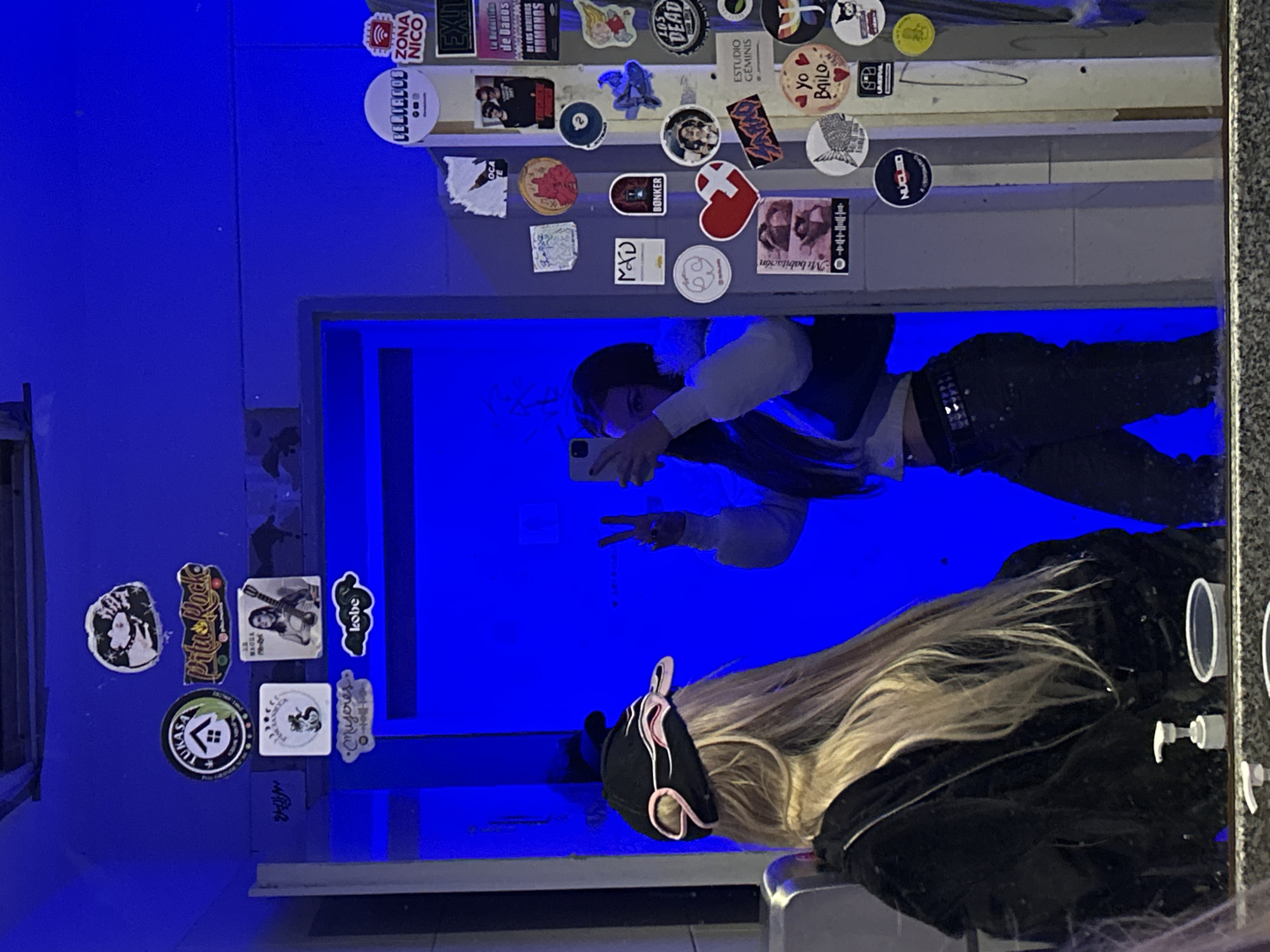 The image depicts a mirror selfie of two people in a bathroom with blue walls. One person is holding a phone and making a peace sign in the reflection, while the other, closer to the camera, has long blonde hair and is wearing a black jacket and hat. The mirror is framed by a collection of stickers, suggesting a public or shared space. The OCR of the stickers is: UKASA, Pitus Racke, MAGRA, mujeres, Kobe, Mi babitación, OCA, CELIACOD ZONA NICO, EX!L, de Danos, DE INS DEREENDS HUMANOS, BONKER, LOS DEAD, ESTUDIO GEMINIS, yo BAILO, NÚCLEO, AMDIRAL. There are visible soap dispensers to the right side. The details suggest a bathroom in a somewhat grungy environment, potentially in the city of Buenos Aires, Argentina, indicated by the Spanish language stickers.