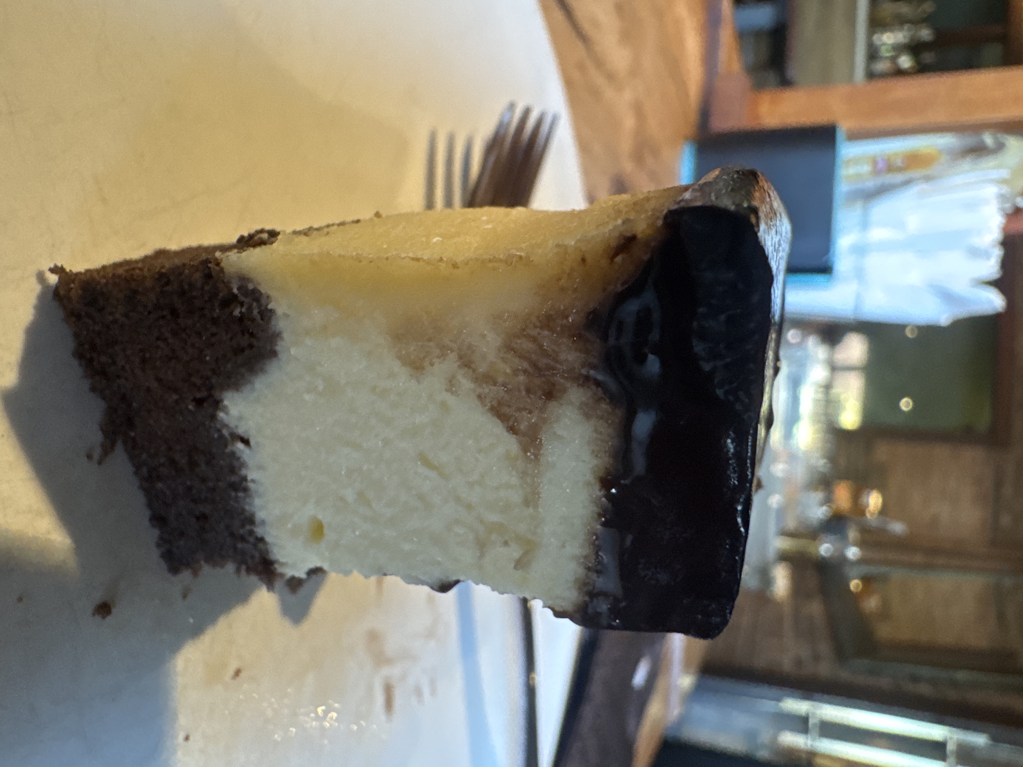 A close-up, eye-level shot shows a slice of chocolate cheesecake on a white plate. The cheesecake has a dark chocolate topping, a thick layer of creamy white filling, and a dark chocolate cake base. A fork is partially visible to the left of the cheesecake, its tines resting on the plate. The background is blurred, showing a rustic interior with wooden elements and what appears to be a bar or restaurant setting. The lighting is warm and natural. The image is static, with no discernible action. The overall impression is of a delicious dessert presented in a casual dining environment, likely in Kuta Selatan, Indonesia, given the context.