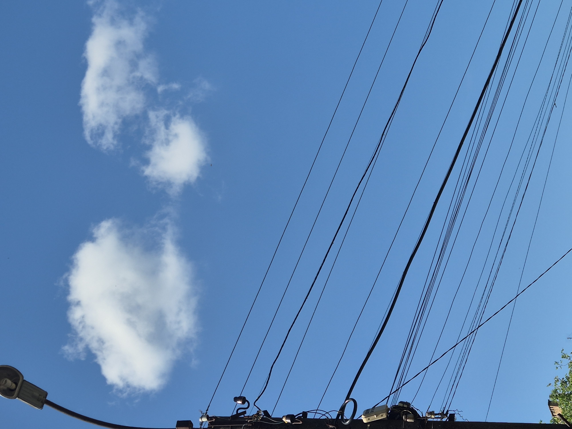 This image, captured in Sozonivka, Ukraine, presents a clear blue sky dotted with two fluffy white clouds. In the foreground, a utility pole is visible on the left side, adorned with multiple black electrical wires stretching horizontally across the frame. A street lamp is mounted at the top of the pole, facing left. The pole itself is cluttered with various electrical components, including insulators and what appears to be a meter box. The bottom left corner of the image shows a glimpse of green foliage and a brick structure, suggesting an urban or suburban setting. The overall scene is a snapshot of an outdoor environment under a bright sky, with a focus on the infrastructure of utility lines.