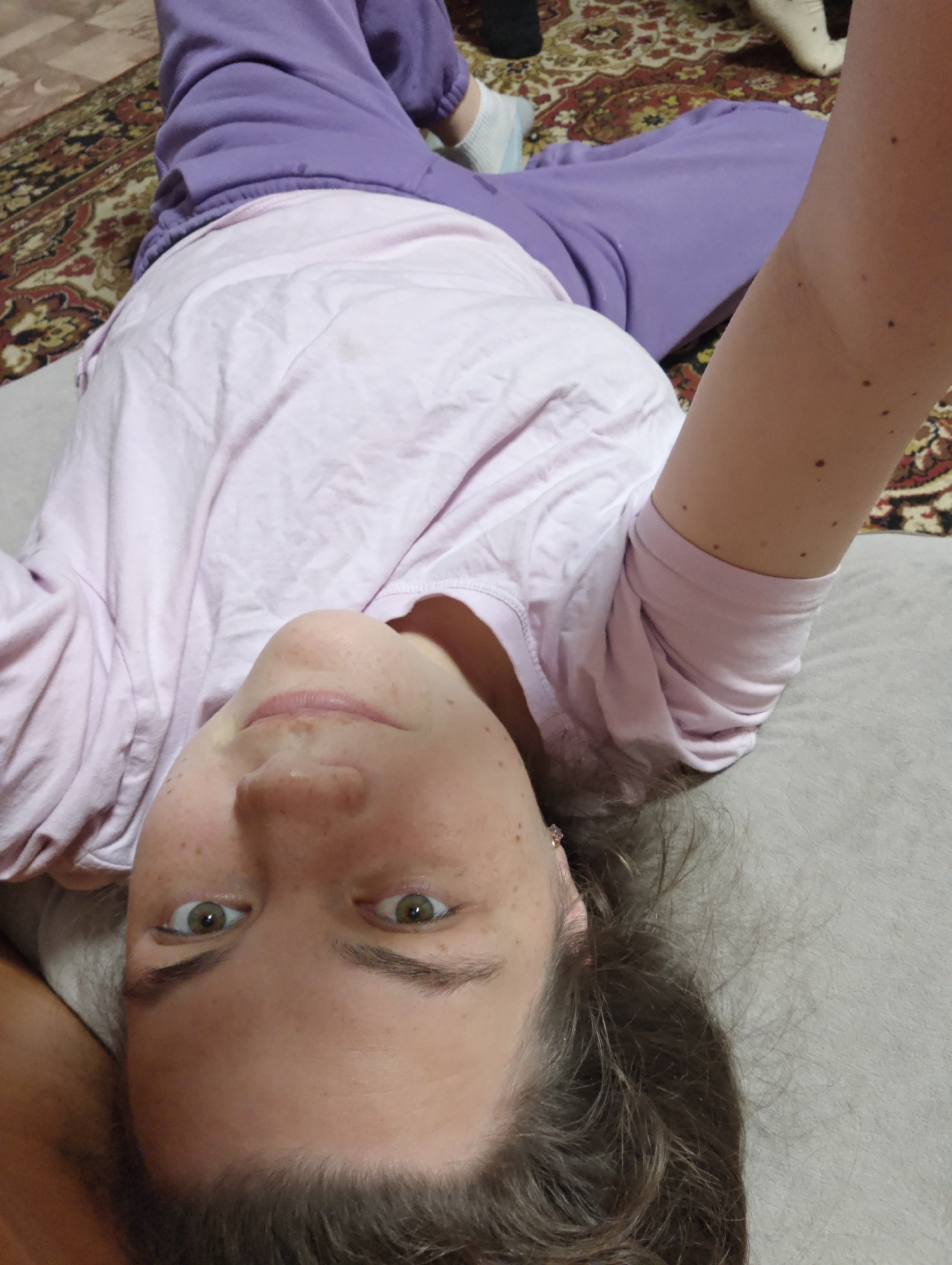 A person lies on a surface, likely a bed or couch, with their head at the bottom of the frame. The person is wearing a light pink t-shirt and lavender sweatpants. Their eyes are open, and they appear to be looking directly at the camera. Some hair is visible at the bottom of the image. A patterned rug is in the background, and the person's arm is raised. It is impossible to verify whether the location of the image is in the city of Уральское, Russia.