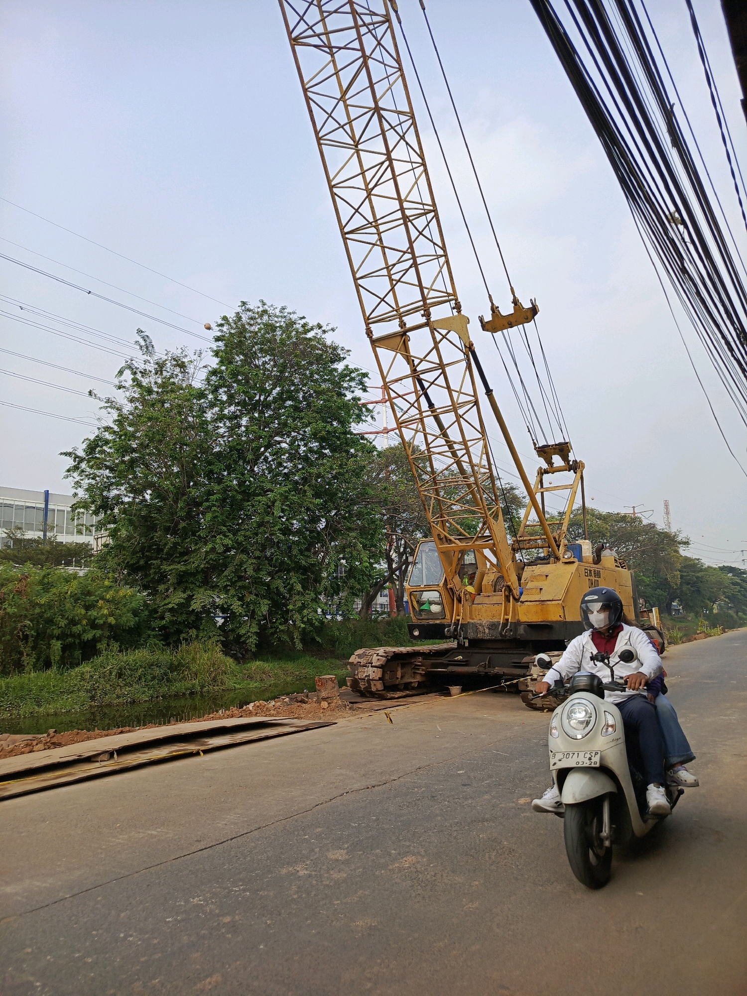In Tangerang, Indonesia, a daylight scene unfolds on a paved road with a large yellow crane dominating the background. The crane, a piece of heavy machinery, has a long lattice boom extending upwards and diagonally across the frame. In the foreground, two individuals are riding on a white scooter. The rider, wearing a white long-sleeved shirt, dark pants, a white helmet, and a white face mask, is operating the scooter. The passenger, behind the rider, is wearing dark blue jeans and white sneakers. The scooter has a license plate that reads "B 3071 CSP". To the left of the road, there is a body of water or ditch with lush green vegetation and trees. In the distance, a building with white and blue accents is visible. Overhead, thick bundles of electrical wires are strung across the sky, supported by poles that are out of frame. The overall environment appears to be an urban or suburban area undergoing construction or development.