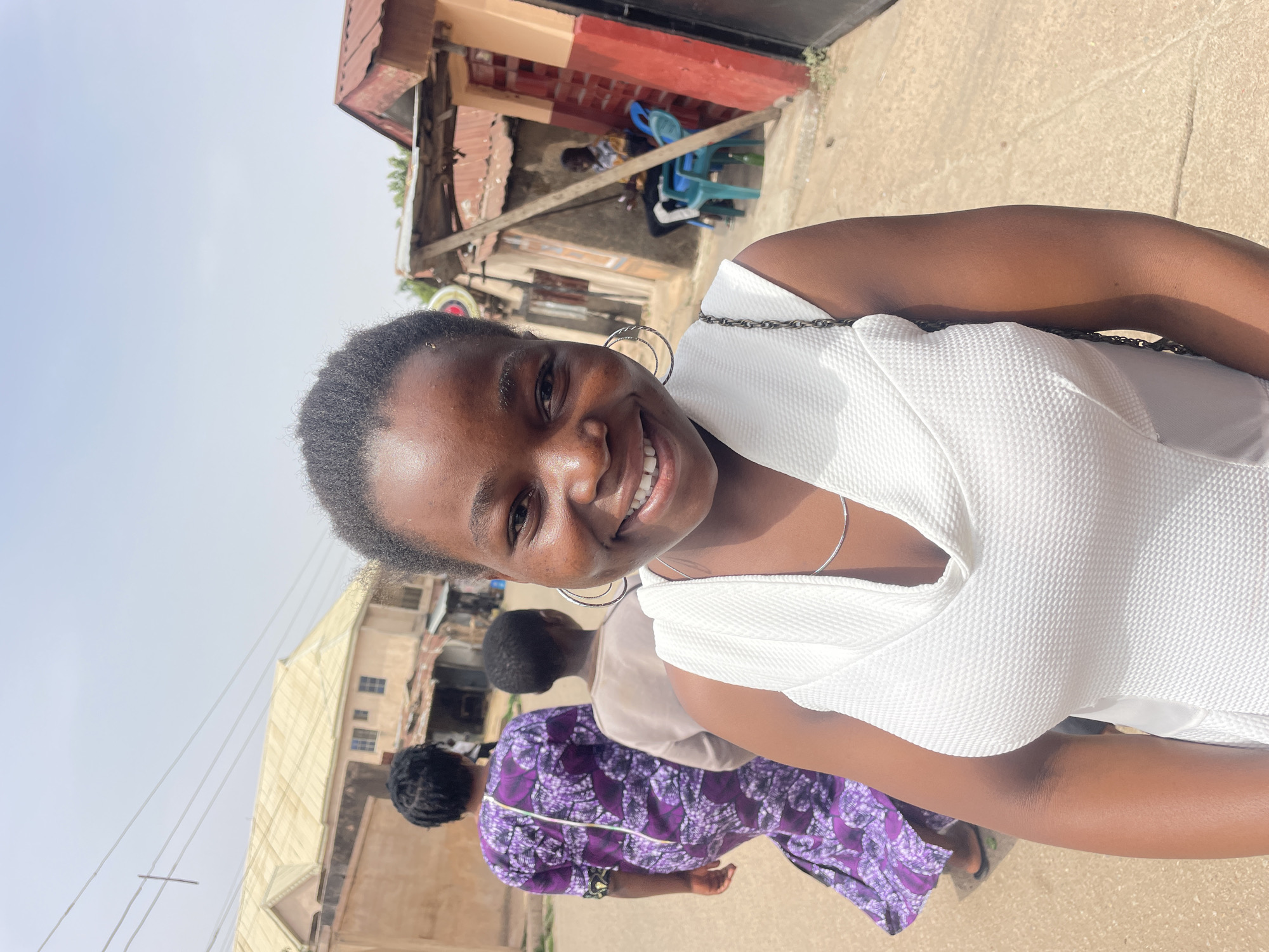 A young woman with dark skin and short black hair is smiling and looking directly at the camera. She is wearing a white top, silver hoop earrings, and a silver necklace. Behind her, two individuals are walking. One is wearing a purple patterned dress, and the other is wearing a tan-colored garment. The background shows a street scene in Jos, Nigeria, with buildings and a partially visible building with a metallic roof visible. In the top center of the frame, there is a building with a red and black exterior, along with blue plastic chairs. The environment appears to be a typical urban area, possibly a residential neighborhood, given the style of the buildings. The photo is taken during daylight hours.