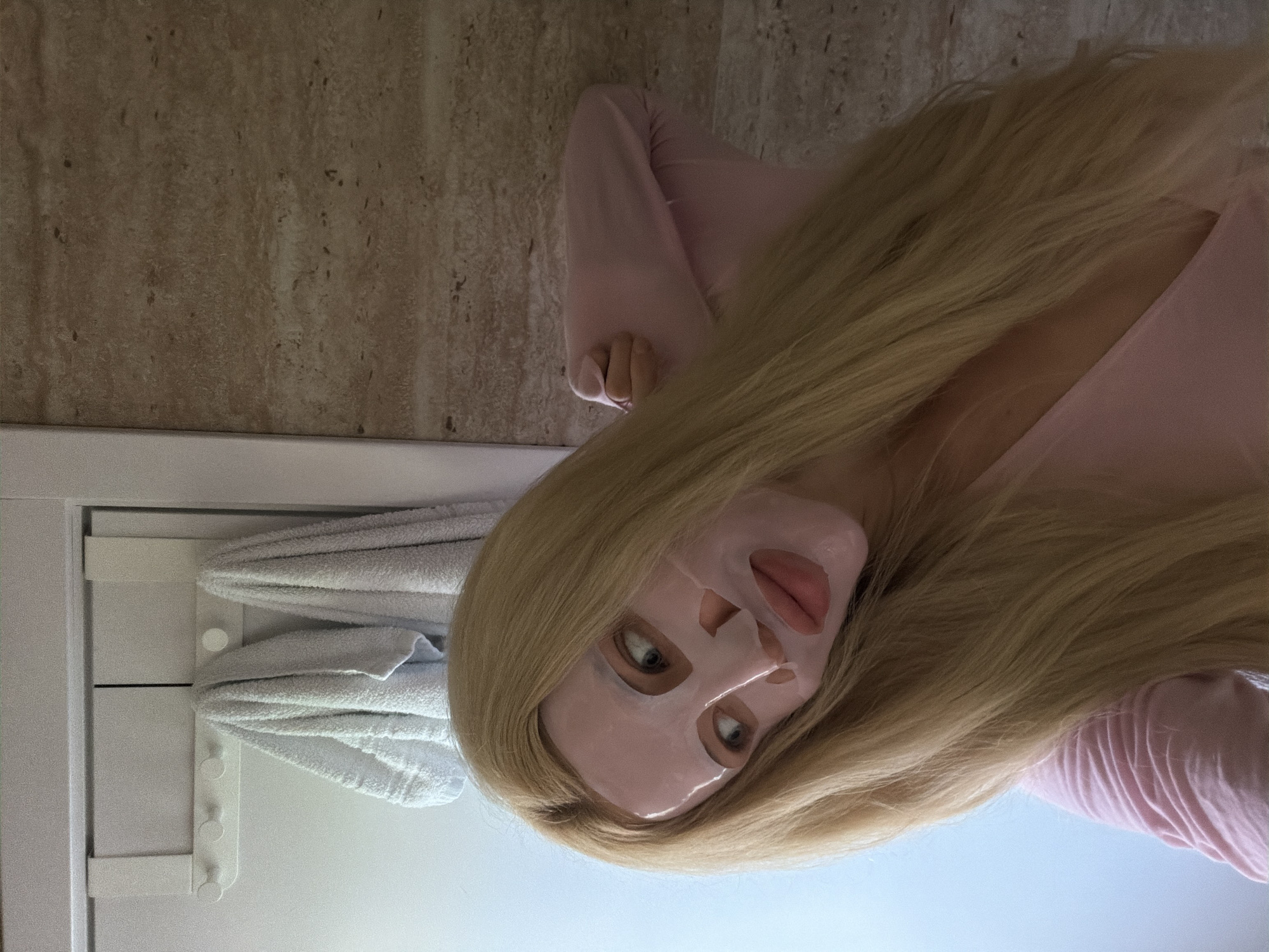 A woman lies on her back in a bathroom with a light pink facial mask. She has long blonde hair and is wearing a pink shirt. A white towel rack with towels is visible. The bathroom has a light-colored tiled wall. The location is potentially Belgrade, Serbia, but there is no definitive evidence of this.