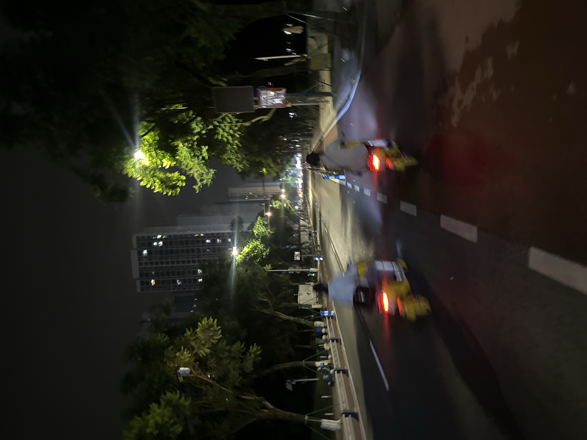 Two individuals are riding electric scooters at night on a paved road in Chongqing High-tech Zone, China. The road is lined with trees and has distinct white lane markings. In the background, tall apartment buildings with illuminated windows are visible. One scooter has a yellow chassis and a red taillight, while the other is less discernible due to motion blur. Both individuals are wearing casual clothing. The scene is illuminated by streetlights and the lights from the buildings.