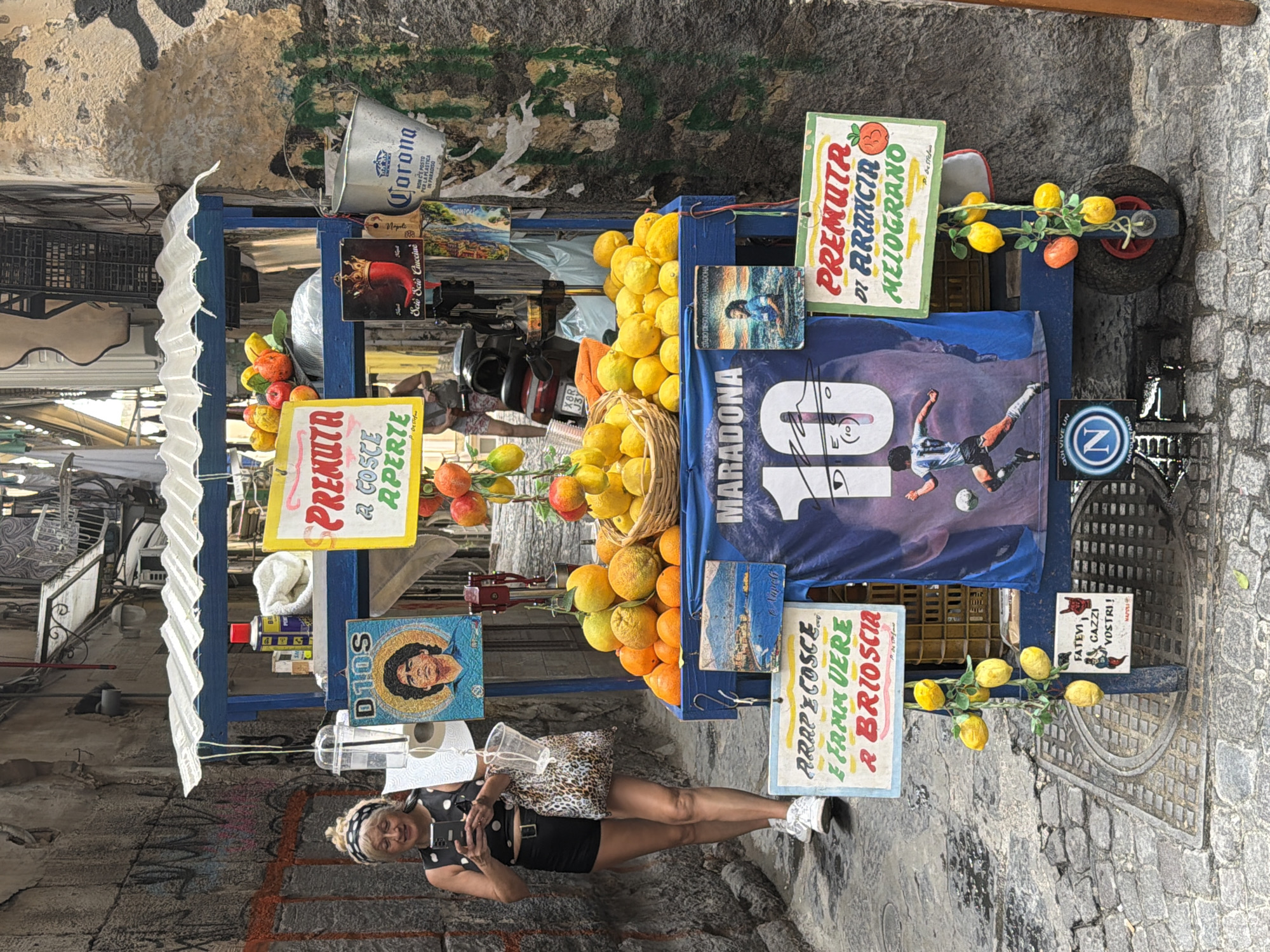 A woman stands in a narrow street in Naples, Italy, looking at her phone. She is wearing a black and white polka dot top and black shorts. To her right, a fruit cart is filled with oranges and lemons. A blue tarp with "MARADONA 10" printed on it is displayed on the cart, along with various signs advertising fresh juice. The street is paved with cobblestones, and the buildings on either side are covered in graffiti and peeling paint.