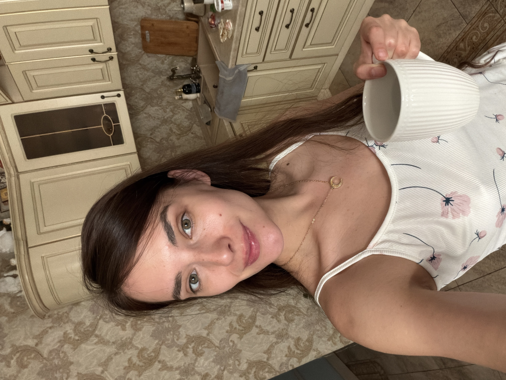 A young woman with long, dark brown hair and green eyes is taking a selfie in a kitchen. She is wearing a white camisole with a floral pattern and a gold necklace with a crescent moon pendant. She is holding a white ribbed mug in her right hand. The kitchen has light-colored cabinets, a light-colored countertop with a sink, and a tiled floor. A wooden cutting board is visible on the counter. The woman is smiling and looking at the camera.