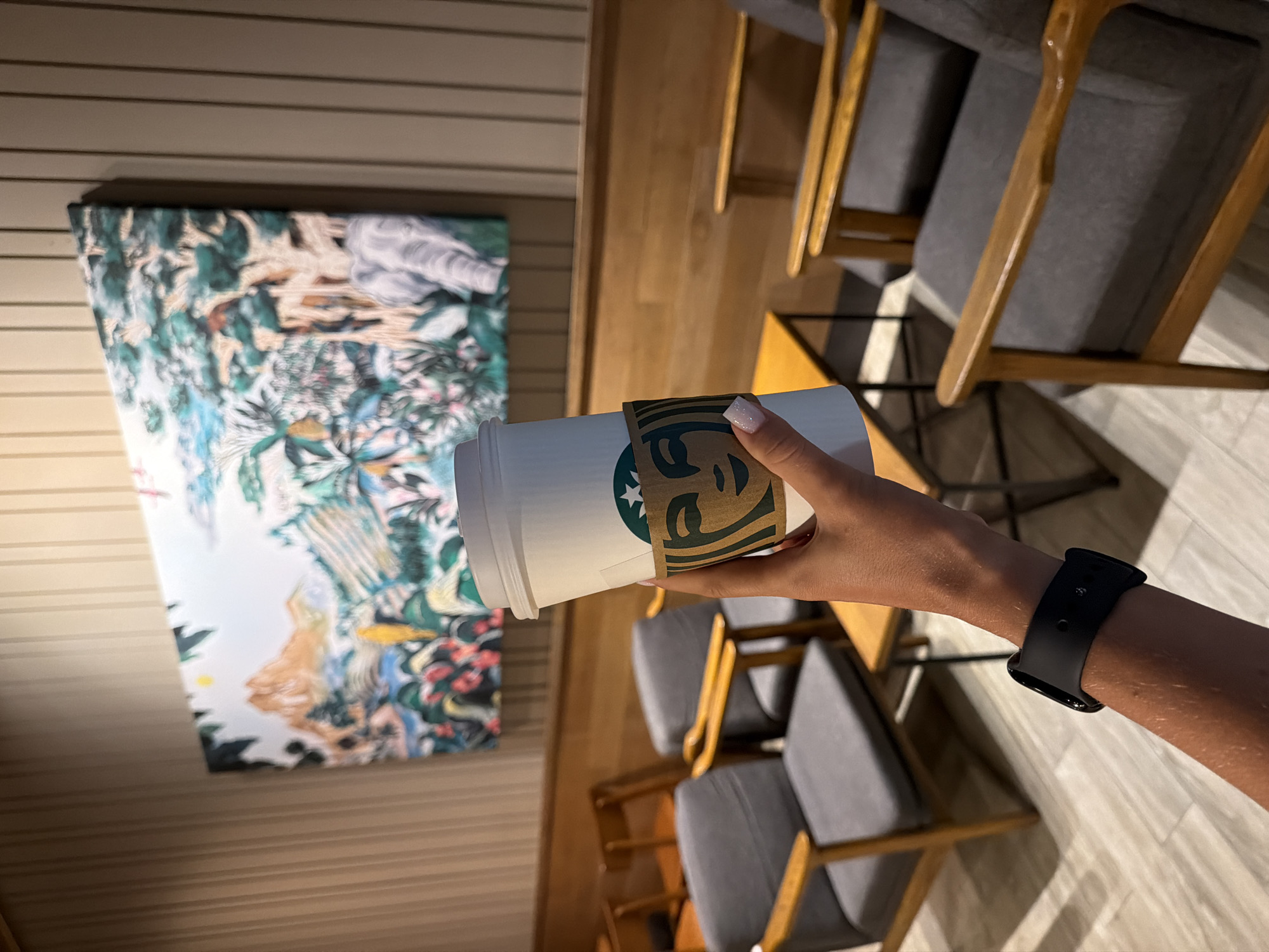 A person holds a Starbucks cup in their right hand, wearing a black smartwatch on their left wrist. Behind the cup are rows of gray and brown chairs and a wall with wood paneling and an abstract painting. The location is Pattaya City, Thailand.